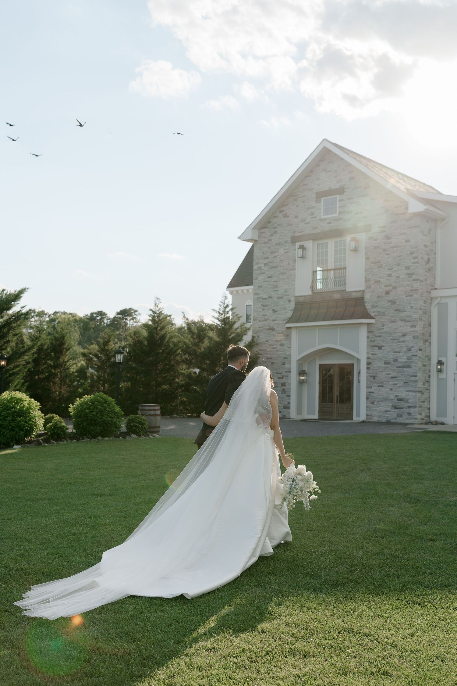 Bride and groom walk toward elegant stone venue as her cathedral veil flows in the breeze.