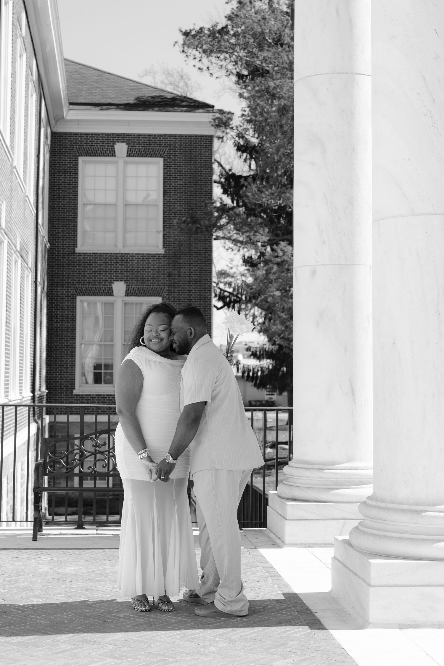 Black and white photo of a couple in white attire sharing a tender moment by marble columns outside a brick building.