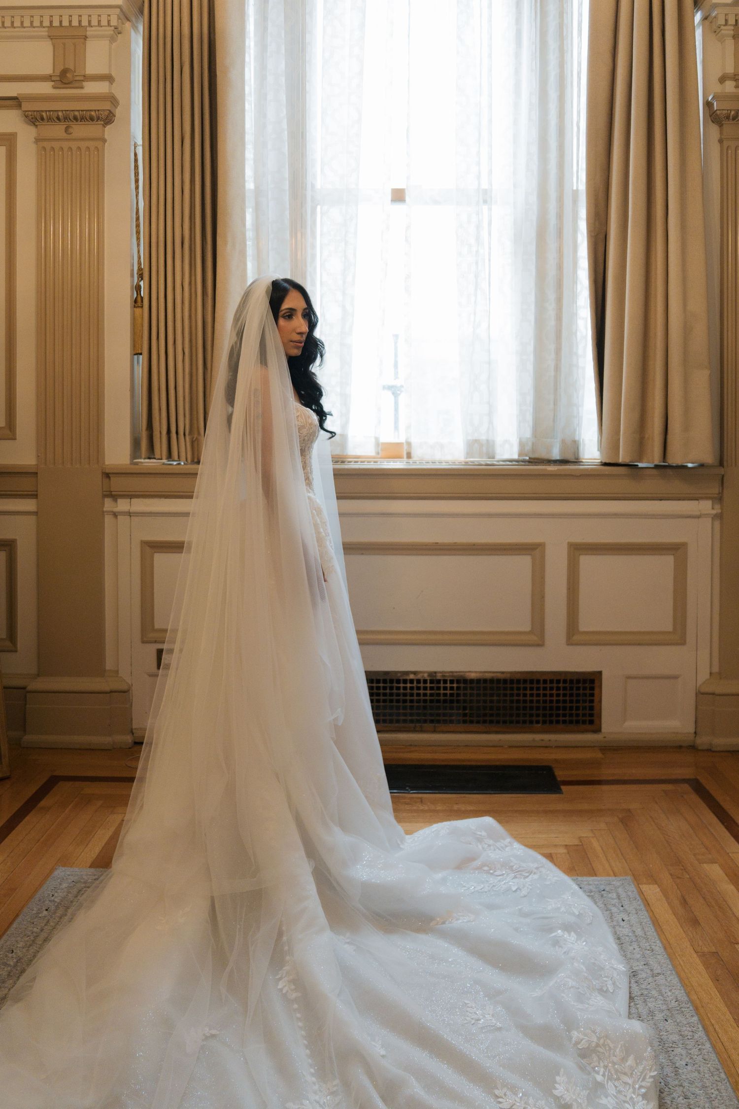 Bride in a stunning white lace wedding gown with cathedral veil standing by an elegant window in a classic venue.