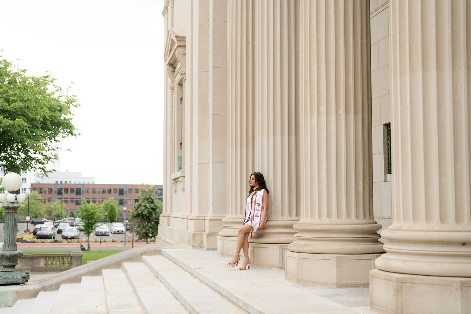 Young woman in striped dress leaning against large stone columns on steps of a neoclassical building.
