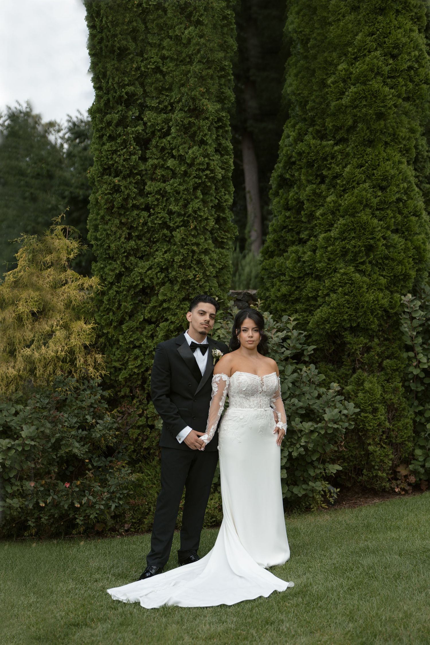 Bride in off-shoulder lace gown and groom in black tuxedo pose together on green lawn before tall evergreen trees.