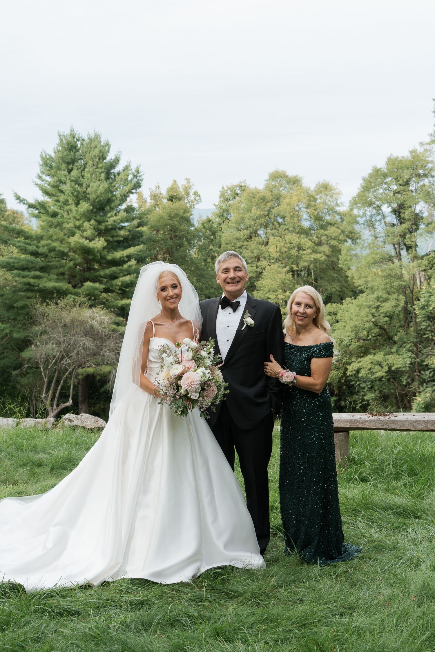 Bride in white gown with bouquet poses with parents in formal attire on a green lawn with trees behind.