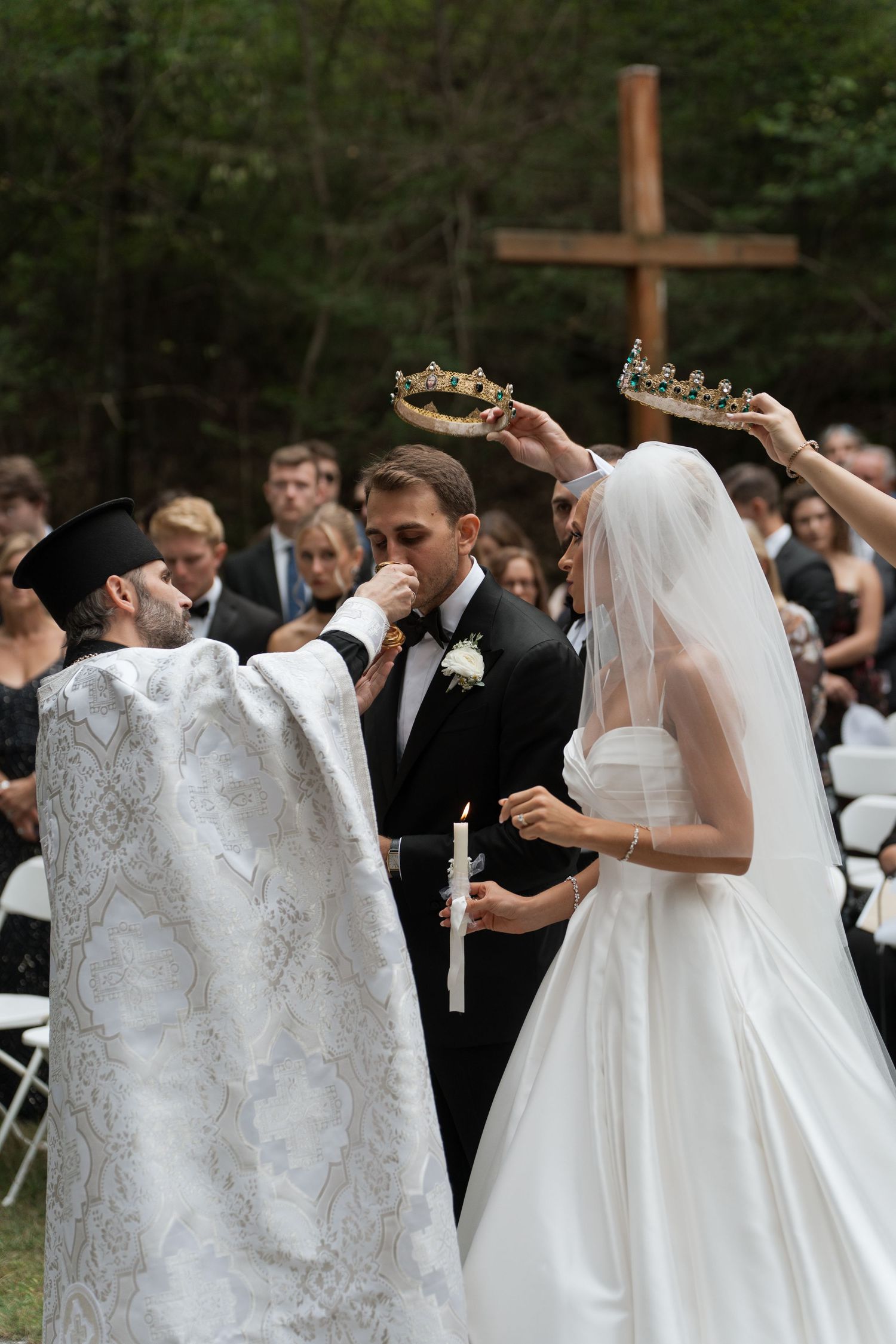 Orthodox priest crowns bride and groom during outdoor wedding ceremony with wooden cross backdrop.