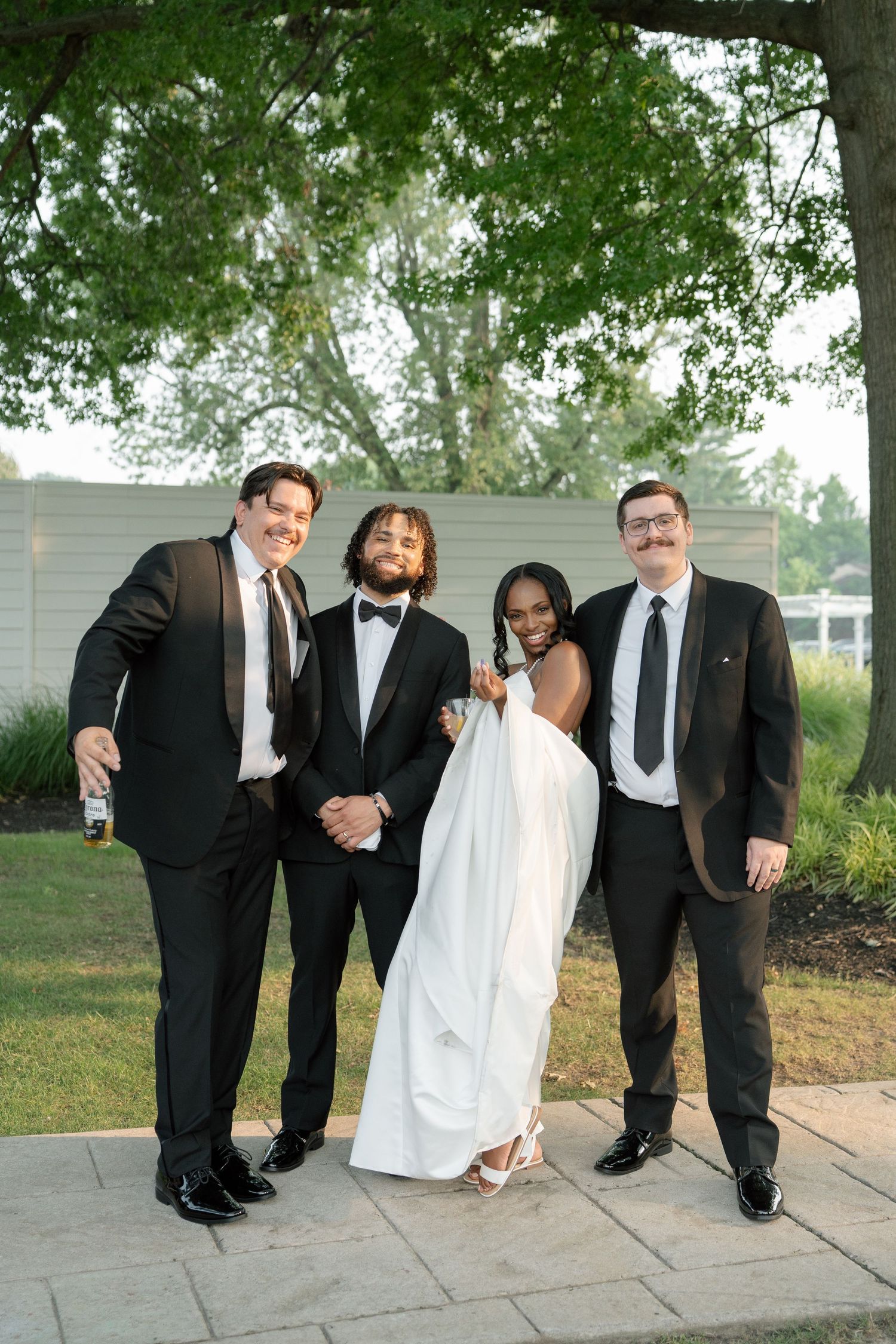 Smiling bride and groom pose with two groomsmen in black tuxedos at an outdoor wedding reception.