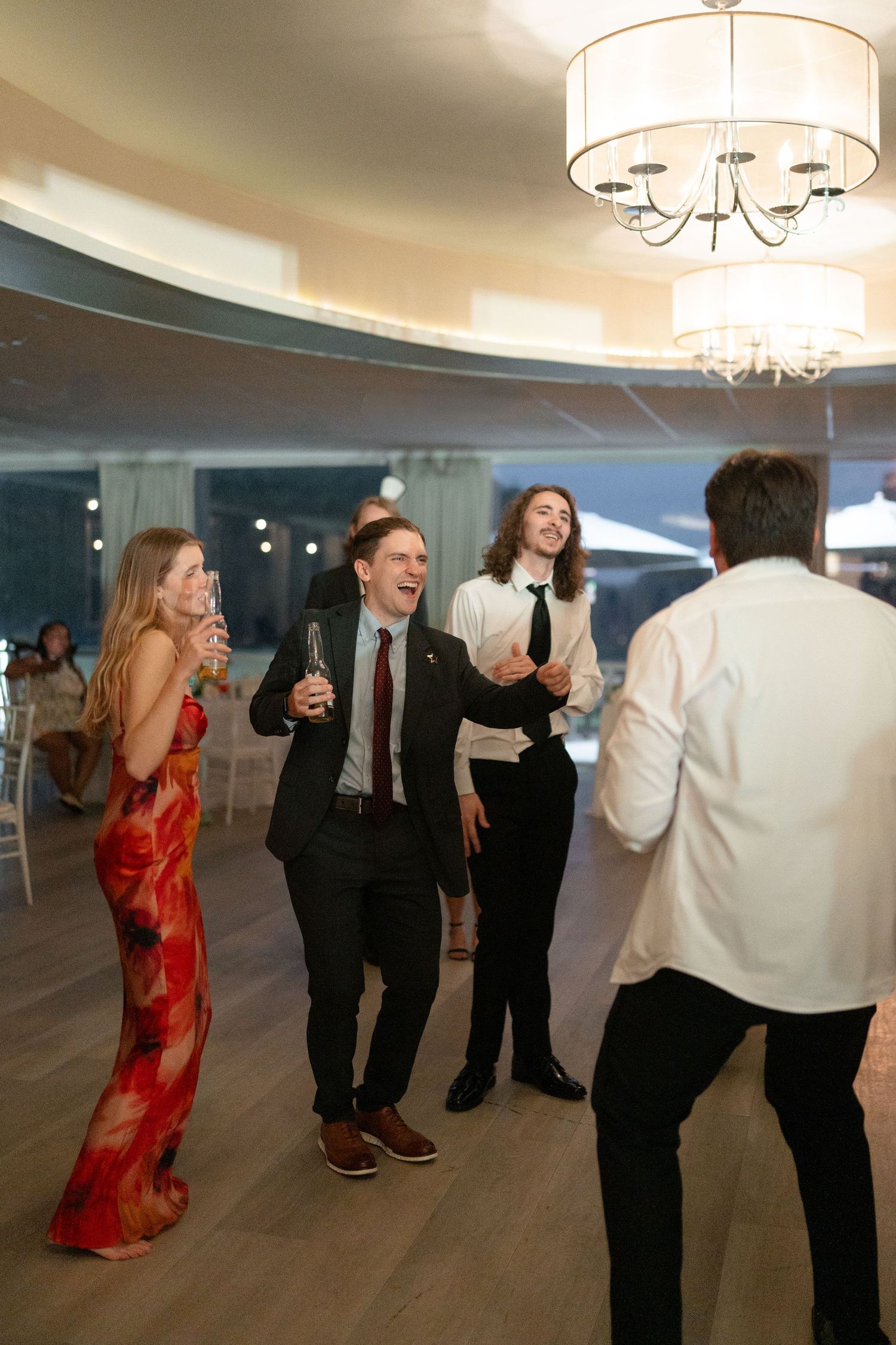 Wedding guests laughing and dancing together under chandelier at elegant reception venue.