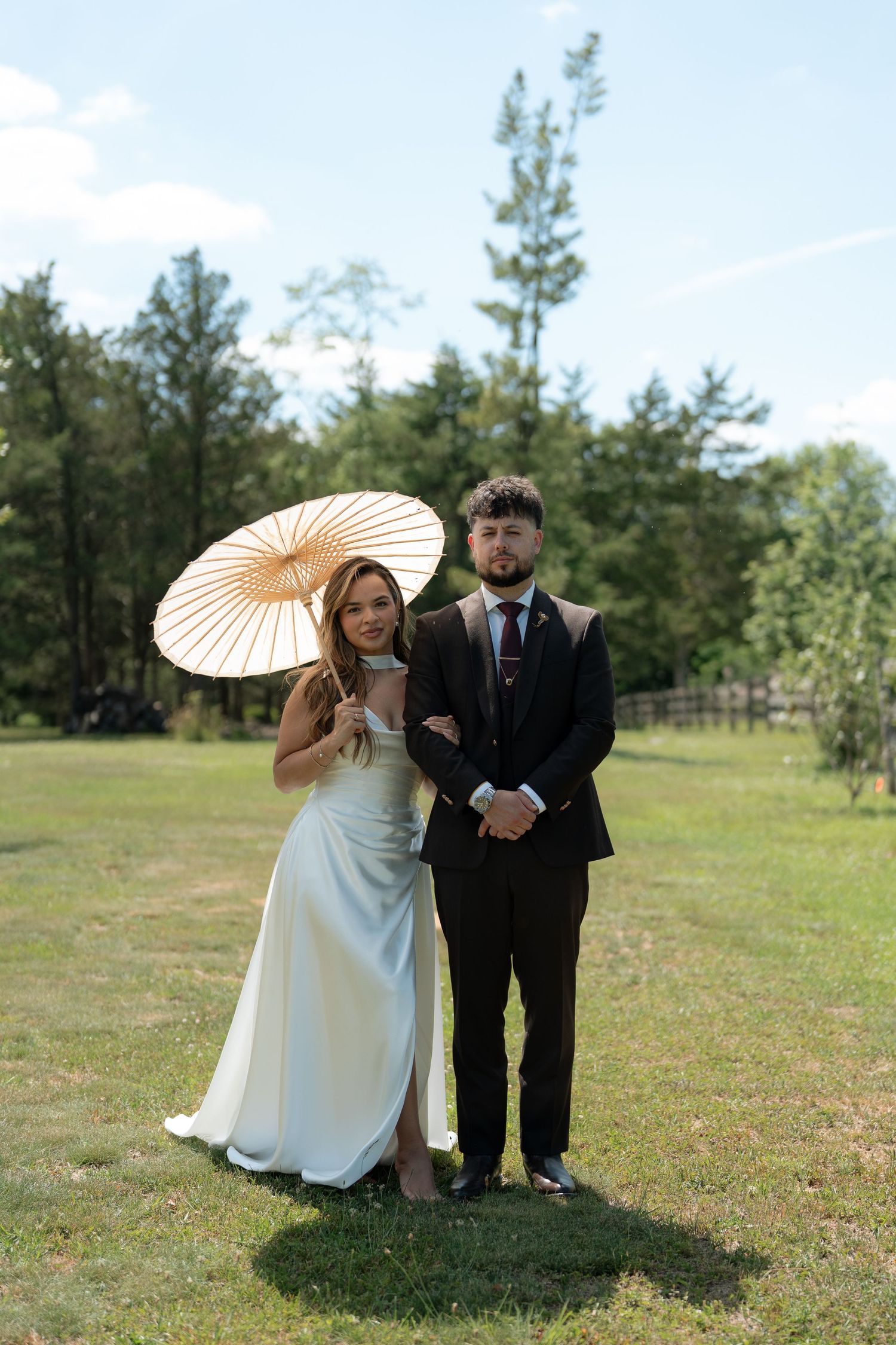 Newlywed couple posing outdoors on green grass, bride in white gown holding parasol, groom in dark suit, trees in background.