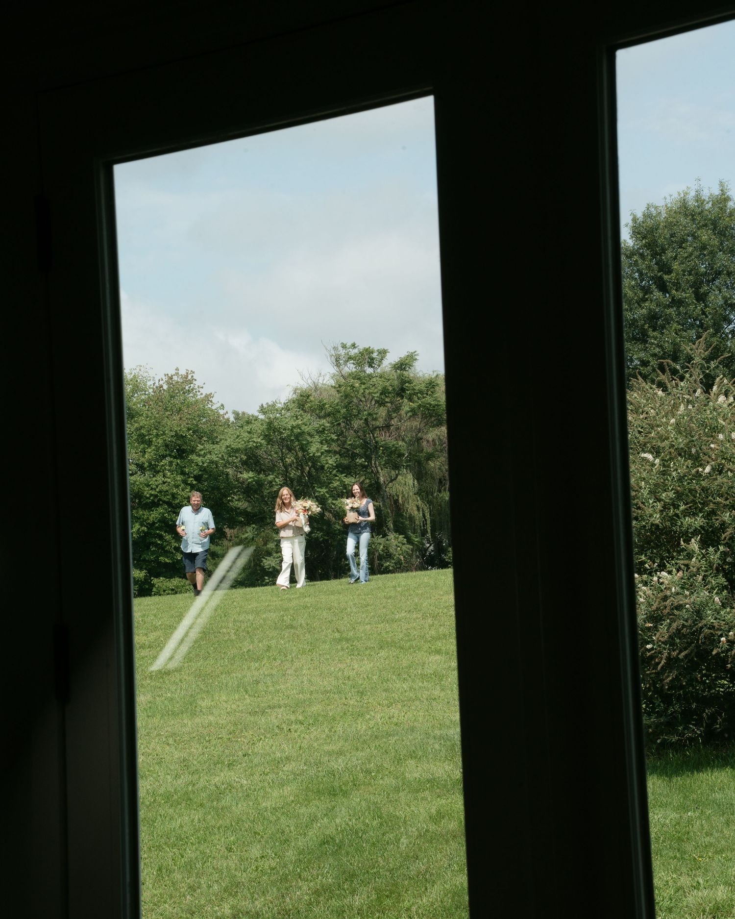 Three people walking across a green lawn viewed through a large window frame on a sunny day.
