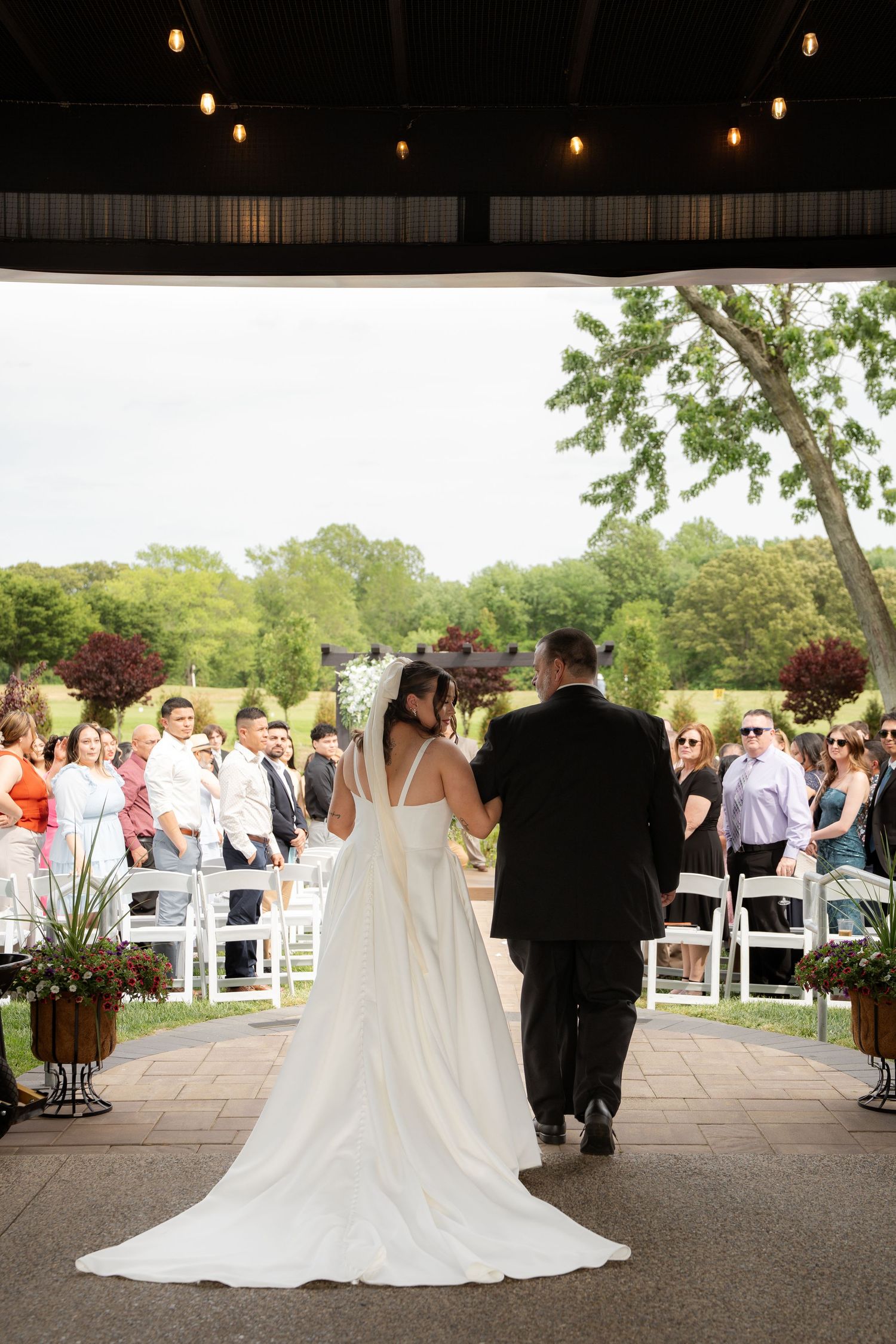 A wedding ceremony with guests watching as a couple walks down an outdoor aisle under string lights.