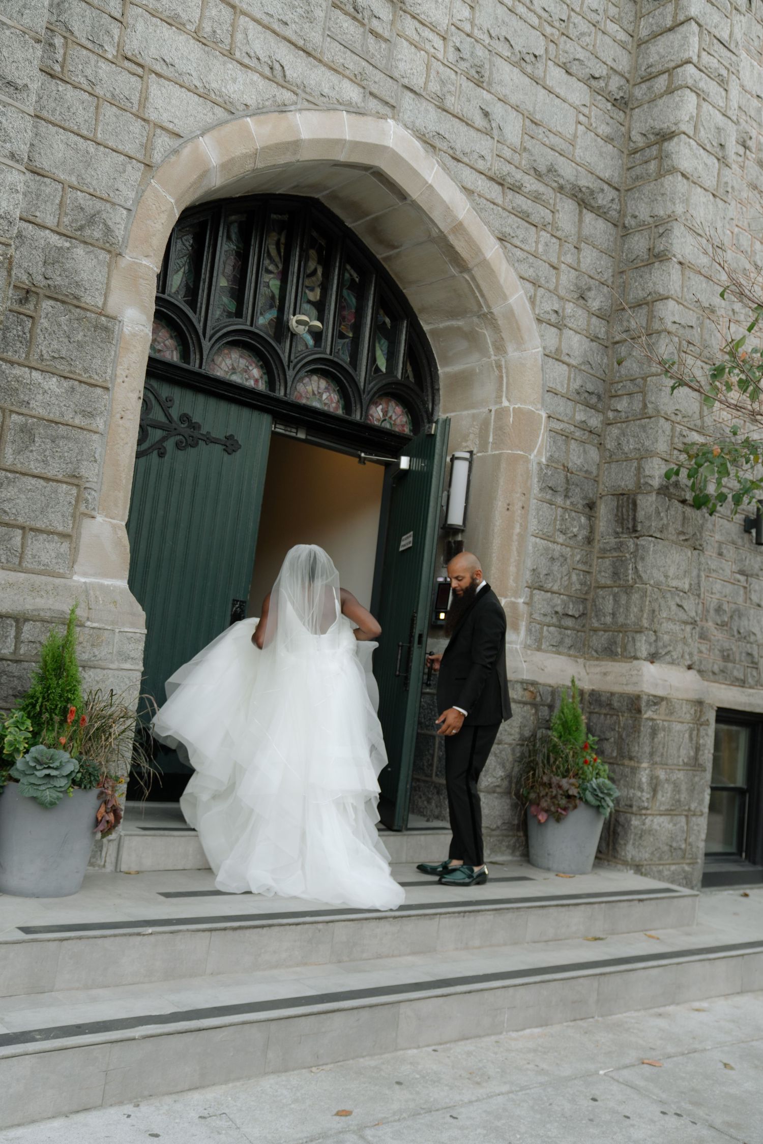 Wedding couple exits stone church building with elegant archway entrance.