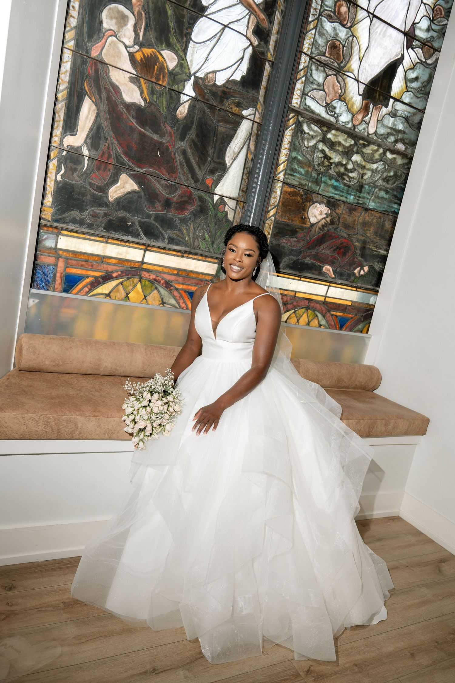 Bride in white ballgown poses in front of ornate stained glass window during wedding photoshoot.