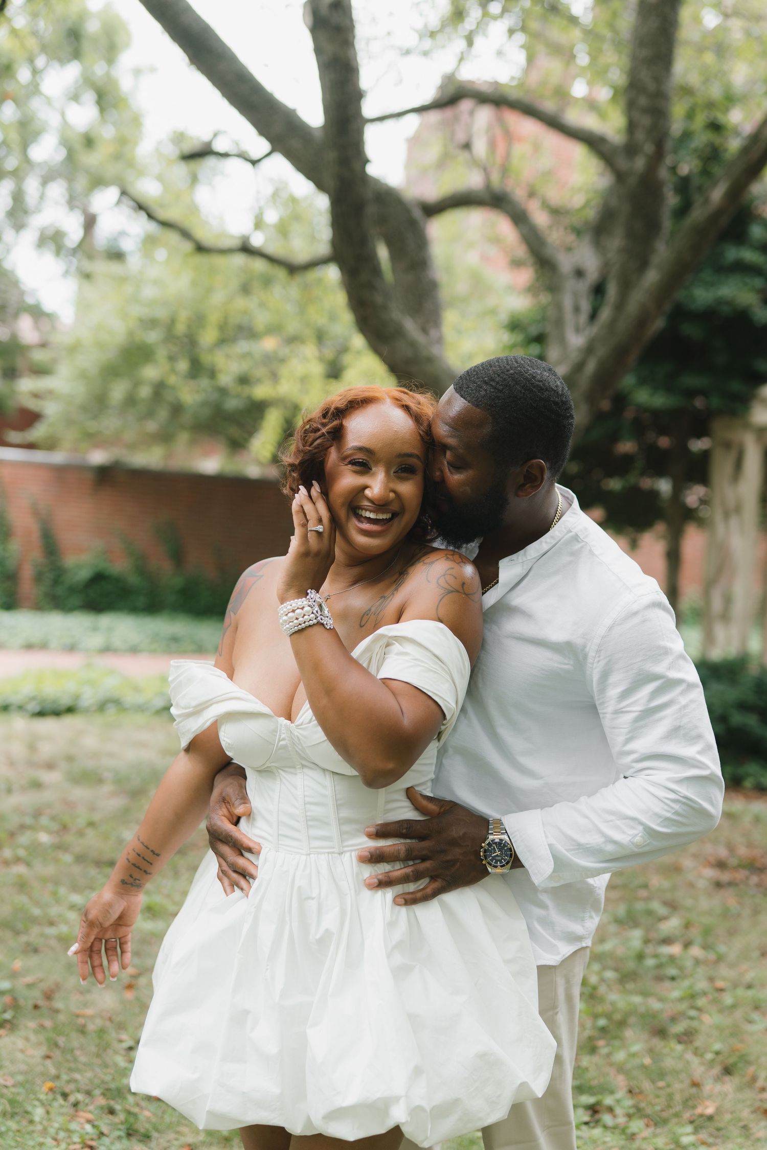 A couple shares joyful embraces and laughter in a park setting while wearing elegant white attire.
