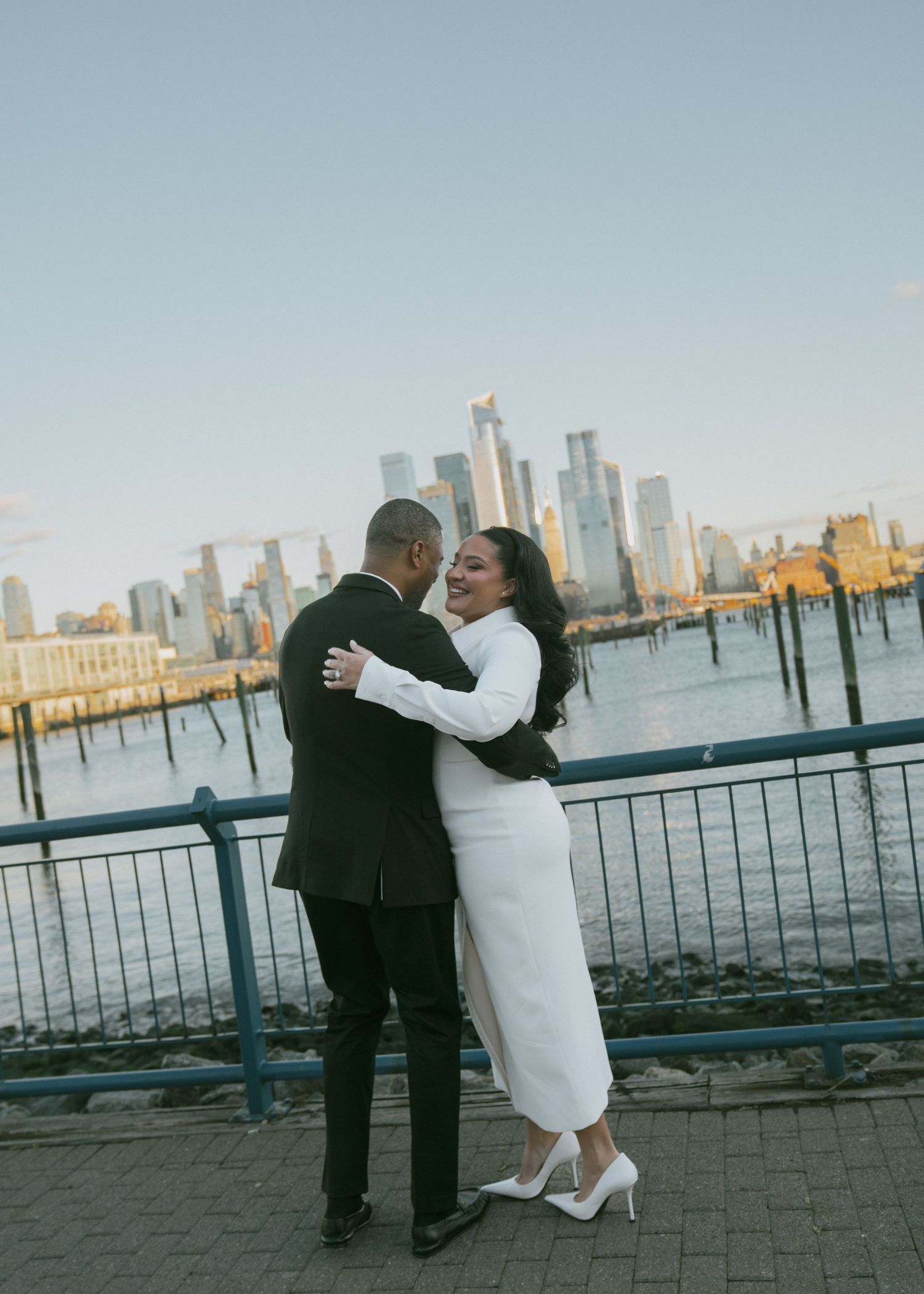 Couple in formal attire posing by waterfront with city skyline in background.