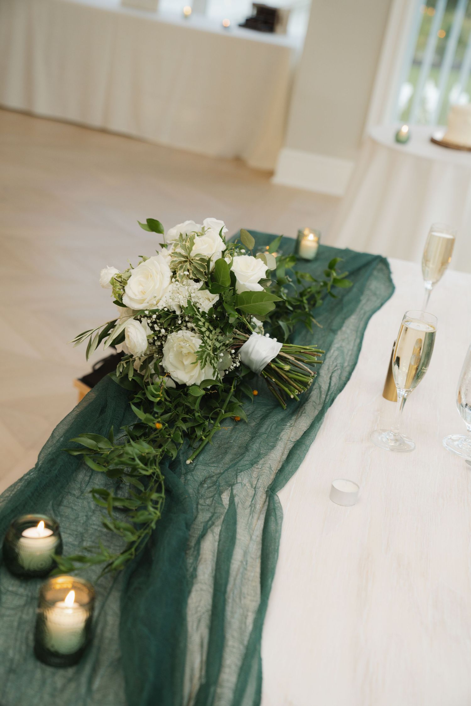 Elegant white roses and greenery arranged on emerald chiffon table runner with glowing candles and champagne flutes.