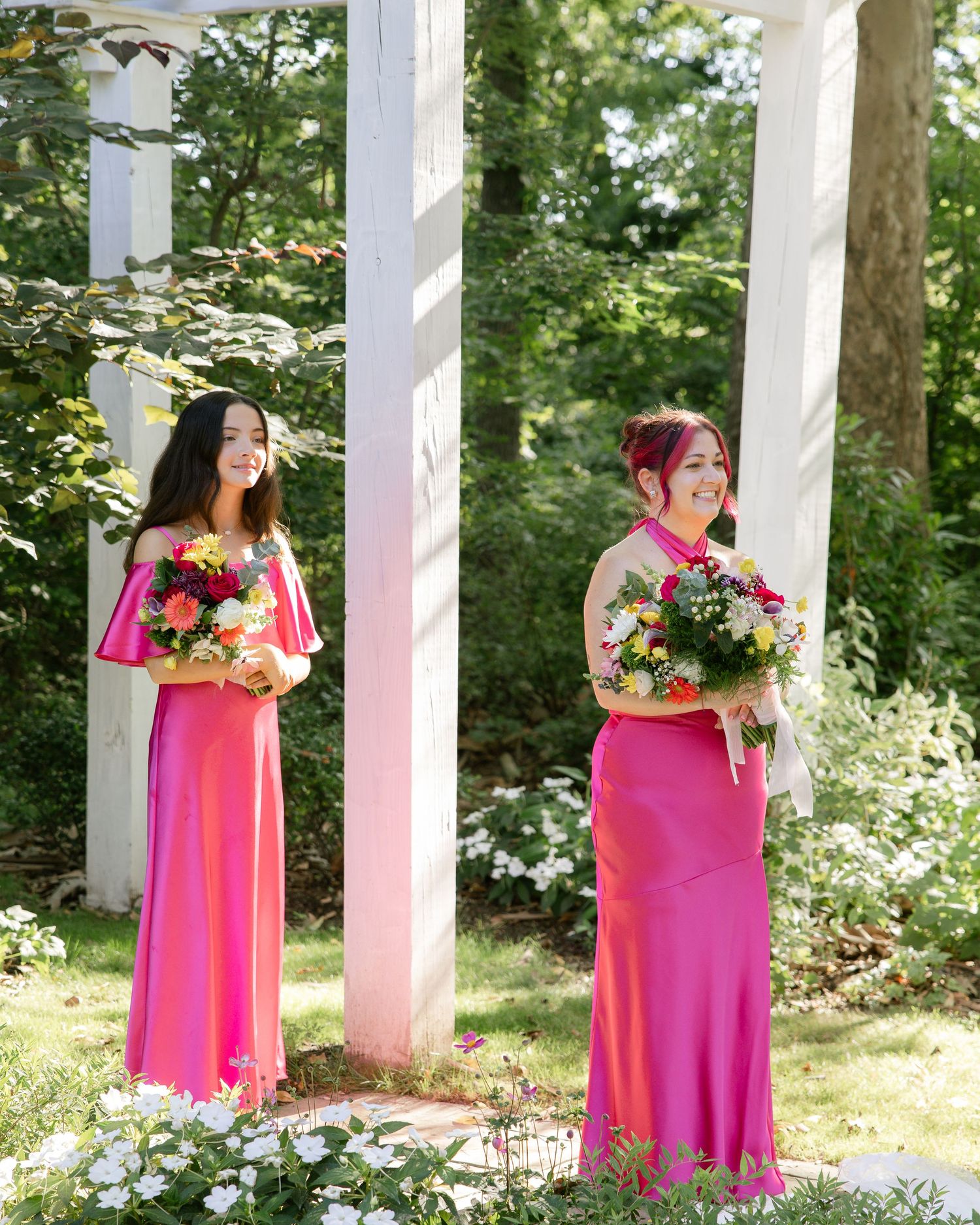 Bridesmaids in bright pink dresses pose with bouquets along white pillars outdoors.