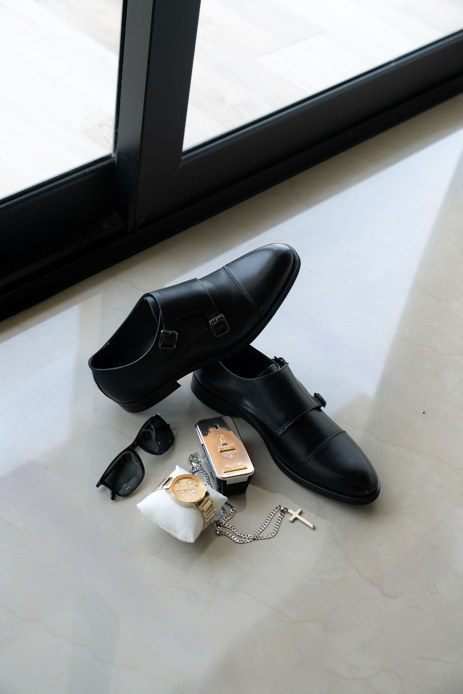 Black leather dress shoes arranged with sunglasses, watch and accessories on white floor near window.
