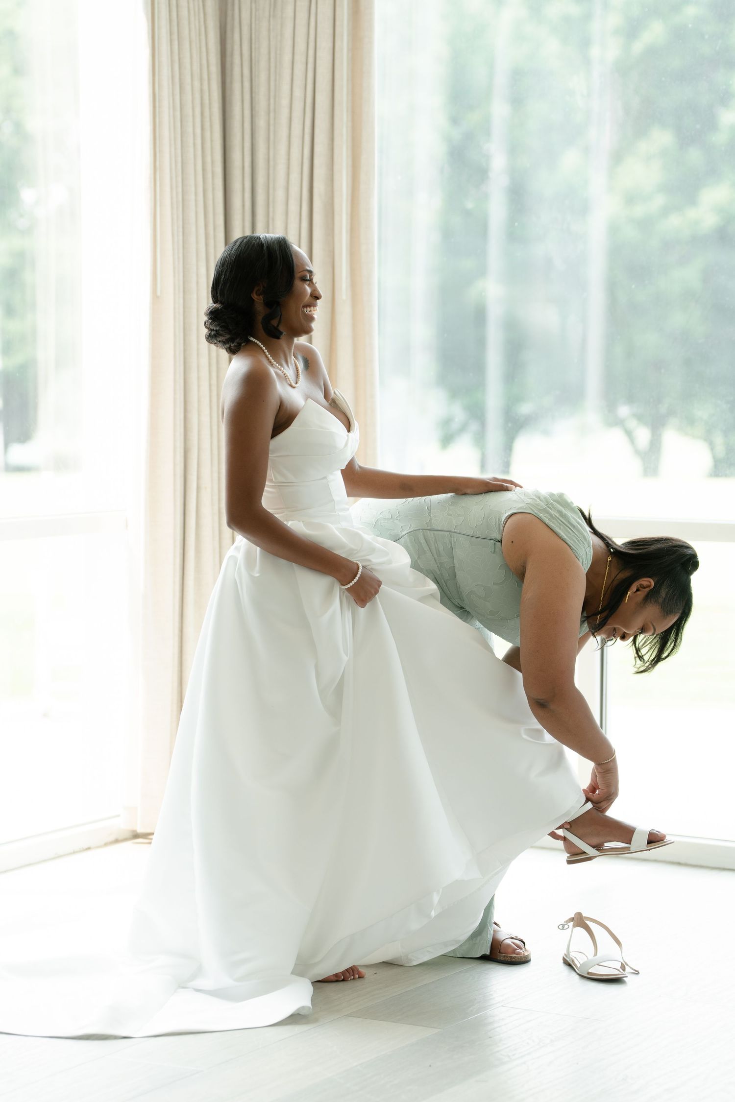 Bride in white gown gets help with strappy heels from bridesmaid in mint dress near bright window.