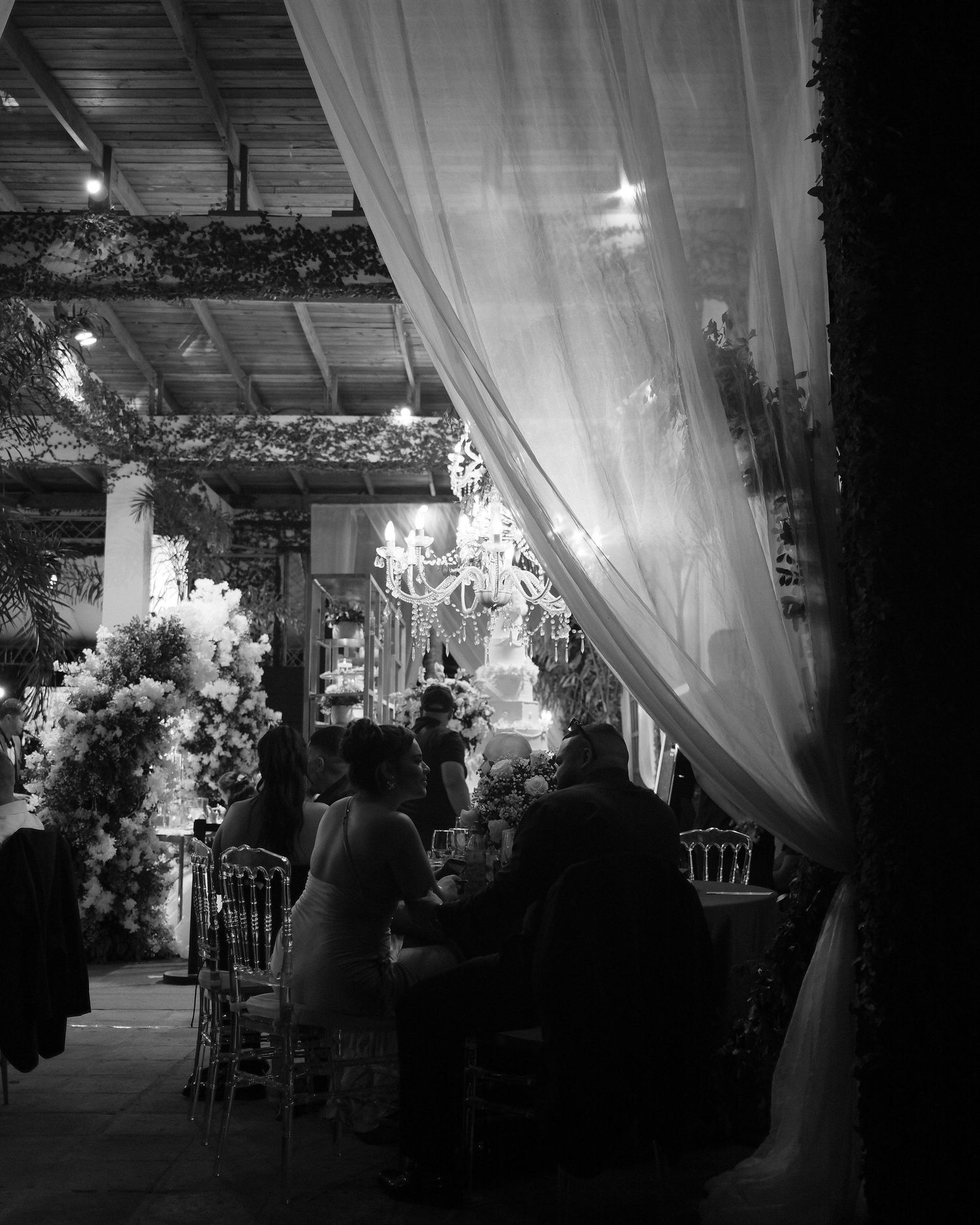 Black and white photo of an elegant wedding reception with floral arrangements, chandelier, draped curtains, and guests seated at tables.