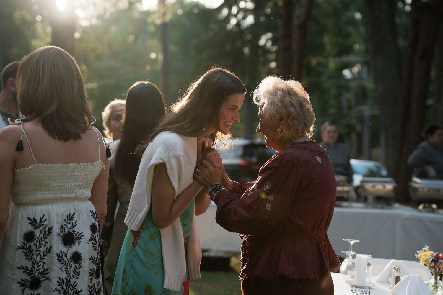 Two women share a warm moment at an outdoor evening gathering, with soft golden sunlight filtering through trees.