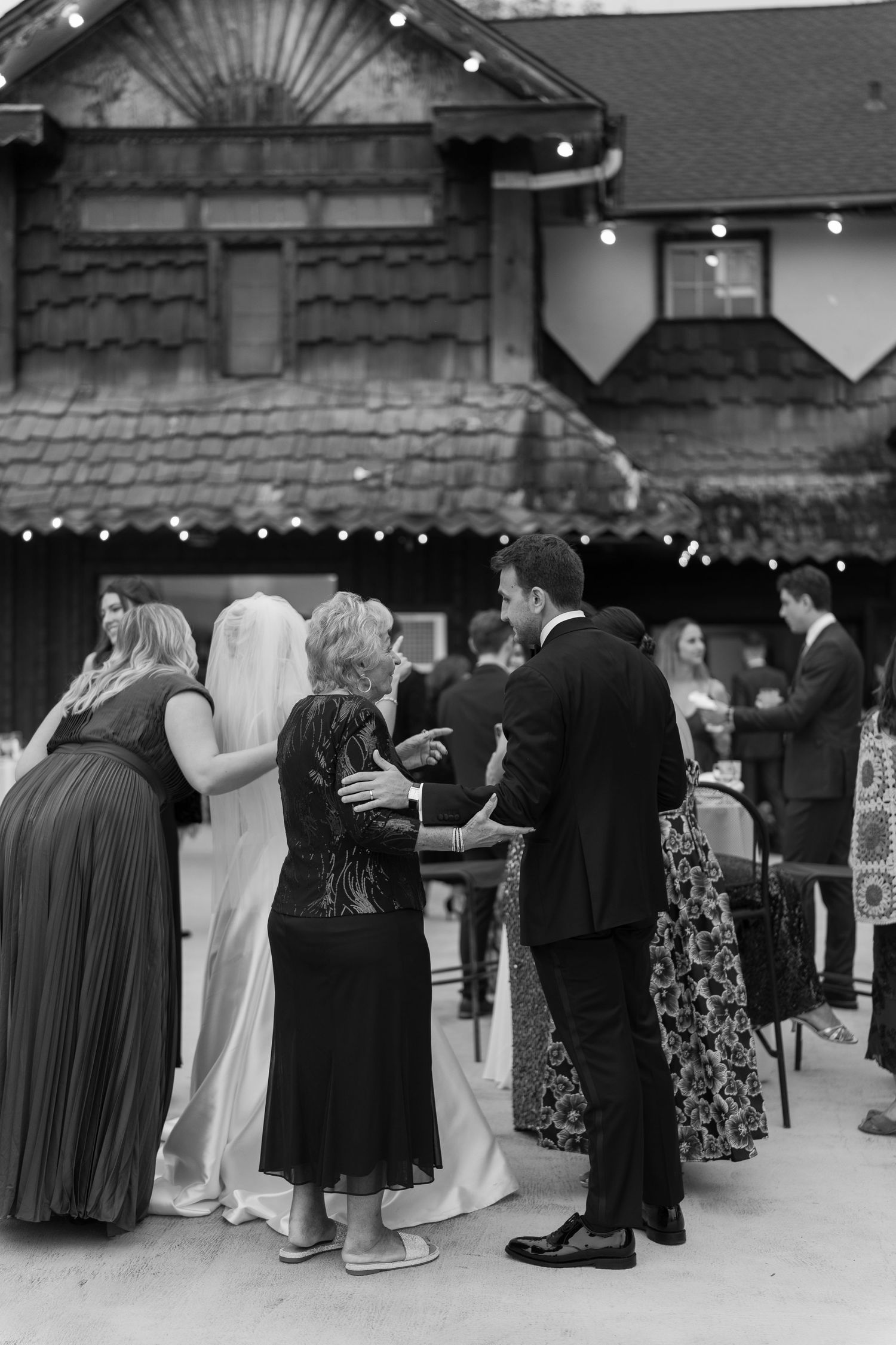 Black and white photo of wedding guests including bride and groom mingling outside rustic venue with string lights.