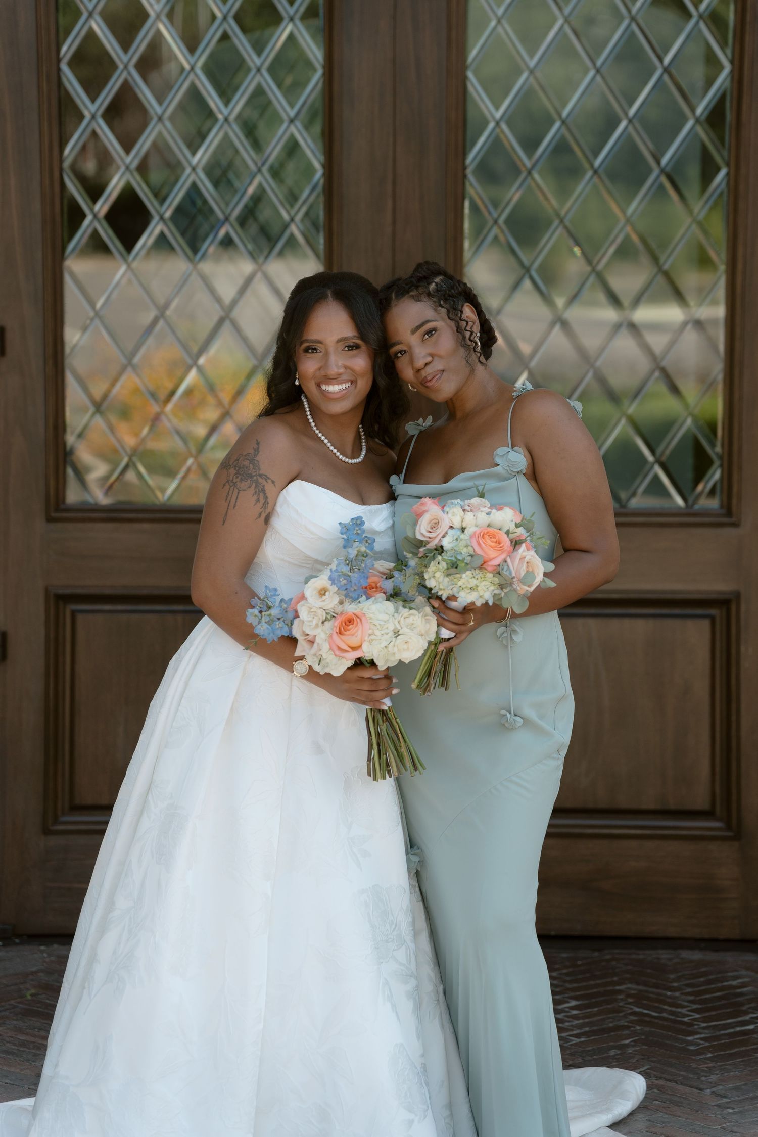 Bride in white strapless gown and bridesmaid in sage green dress holding bouquets of peach and white roses.