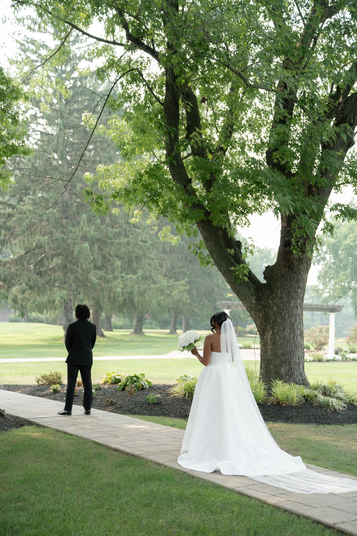 Bride in white gown holding bouquet approaches groom on garden path beneath lush green trees.