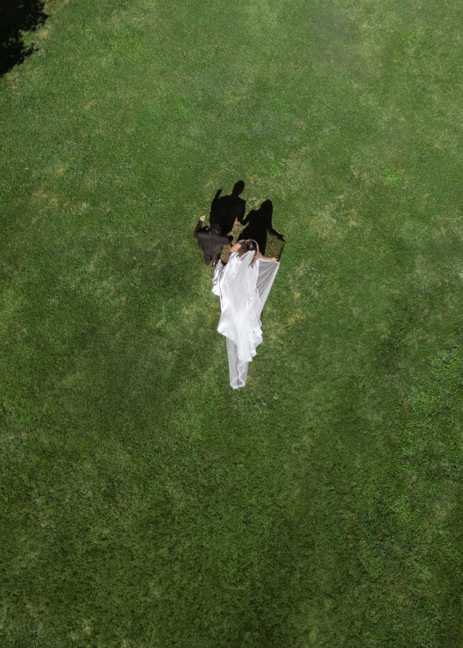 An aerial drone view of a bride in a white gown and veil standing with her groom on a lush green lawn.