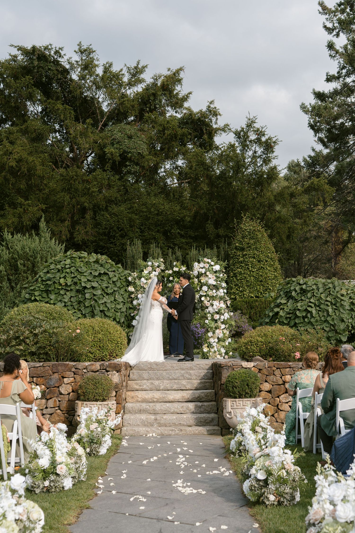 Bride and groom exchange vows at an elegant outdoor garden ceremony with floral arch and white petal-lined aisle.