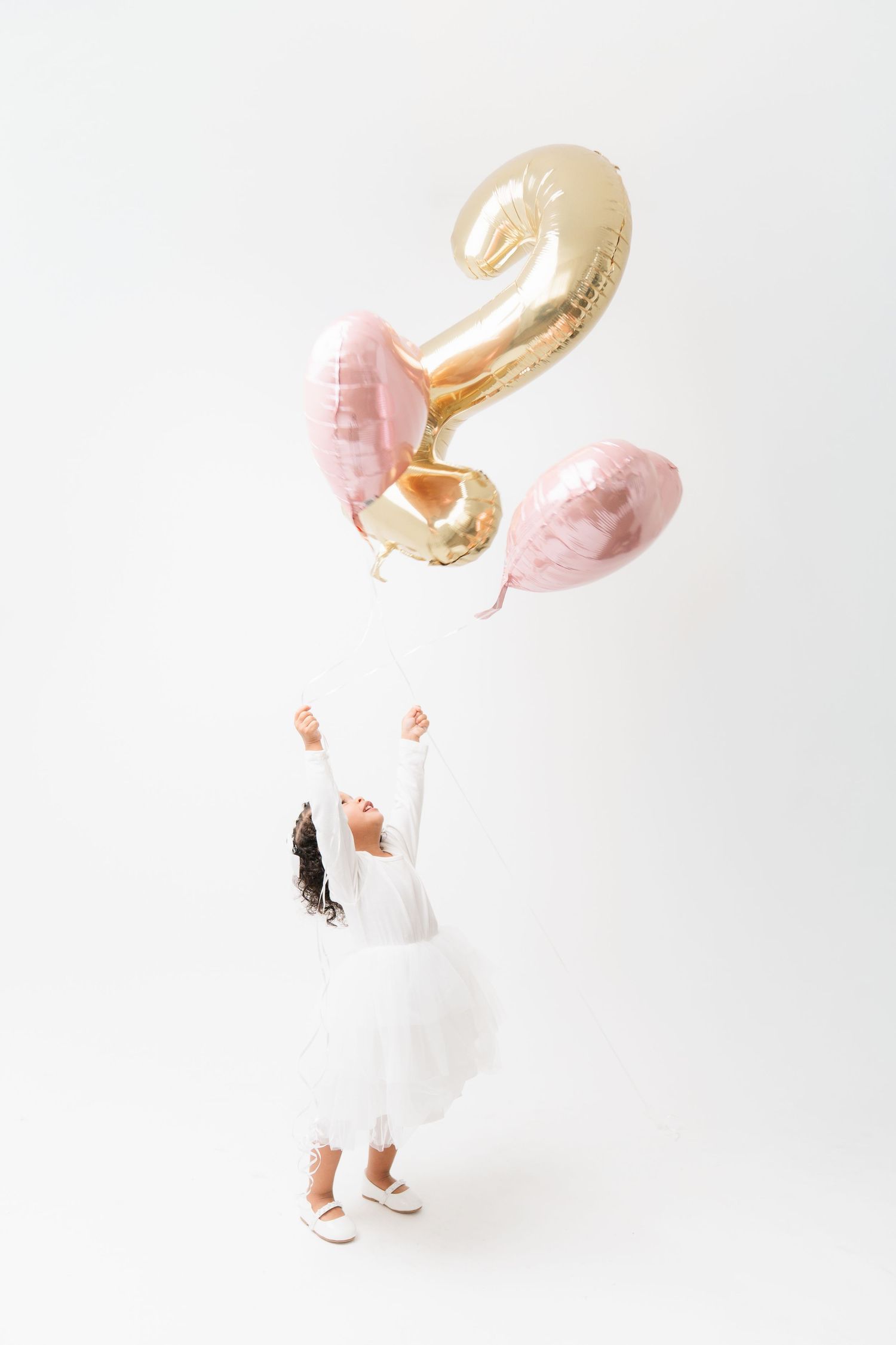 Child in white outfit playing with gold balloons against white background.