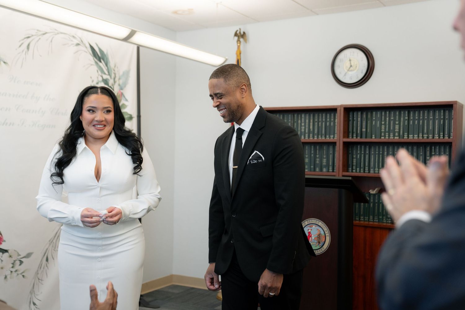 A couple in formal attire stands together in an office setting with bookshelves.