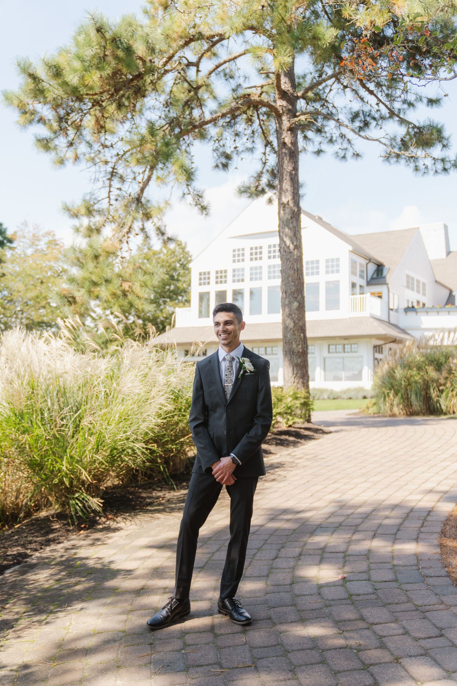 A groom in a classic black suit poses on a curved brick pathway surrounded by pine trees and sunlight.