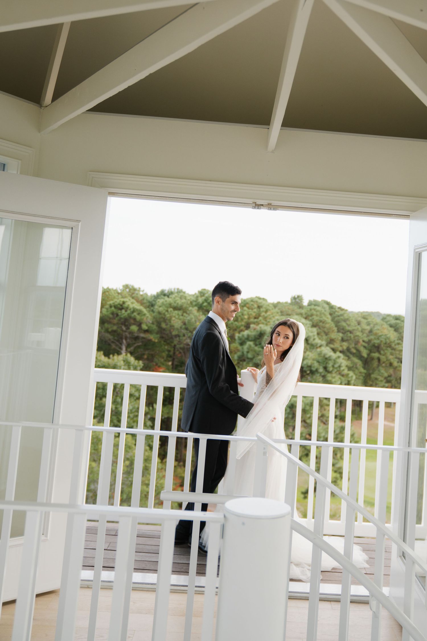 A bride and groom share playful poses along a white gazebo railing with scenic greenery in the background.