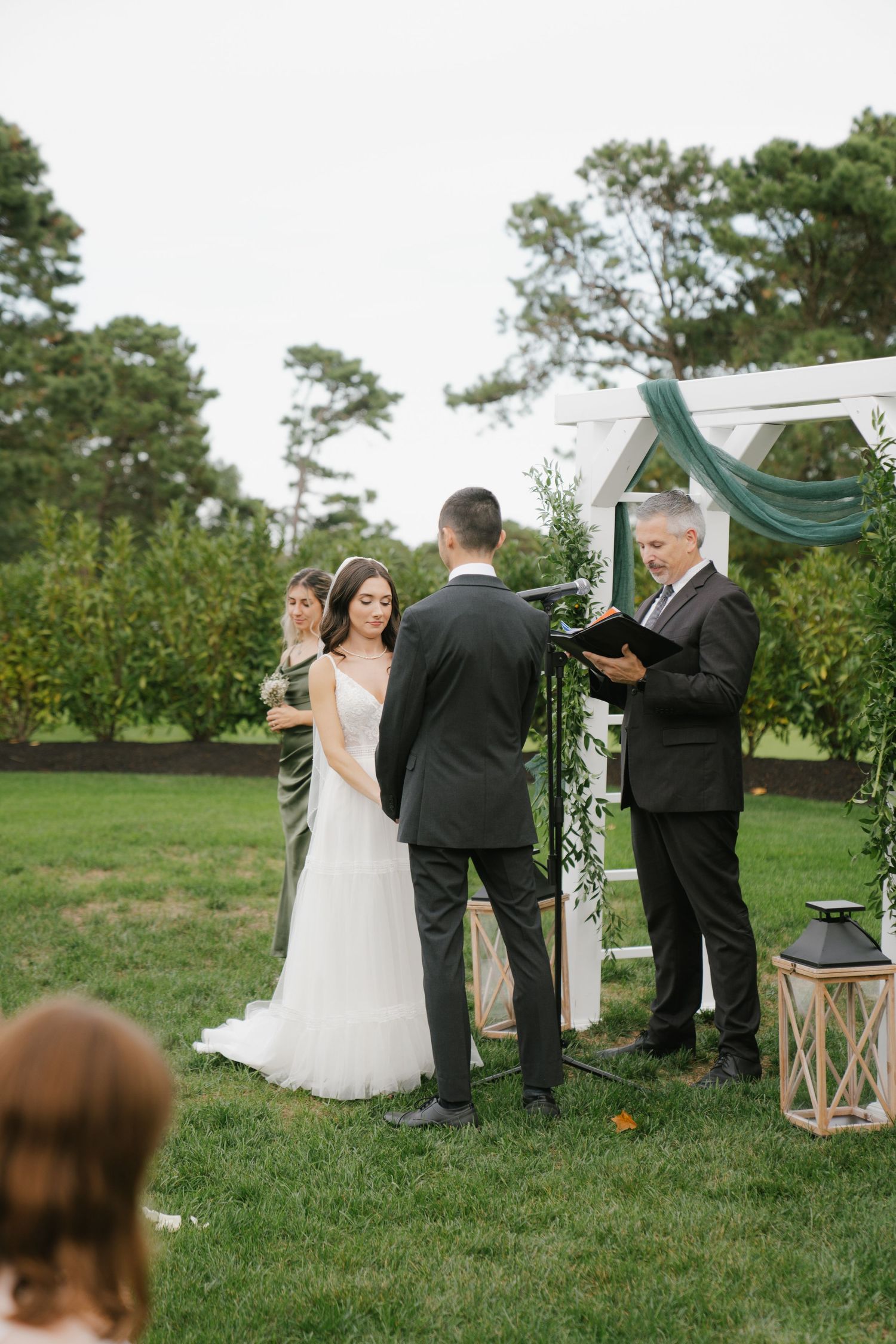 An outdoor wedding ceremony takes place under a white arch decorated with teal fabric and greenery in a garden setting.