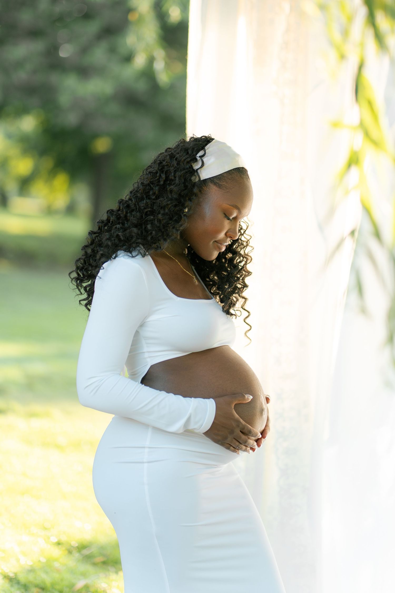 Pregnant Black woman in white dress cradling baby bump outdoors near sheer curtains in sunlit park setting.