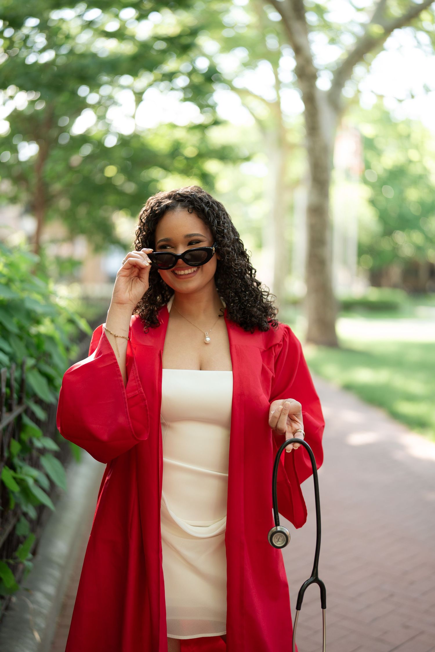 Young woman in red graduation gown holding stethoscope and adjusting sunglasses in a tree-lined walkway.