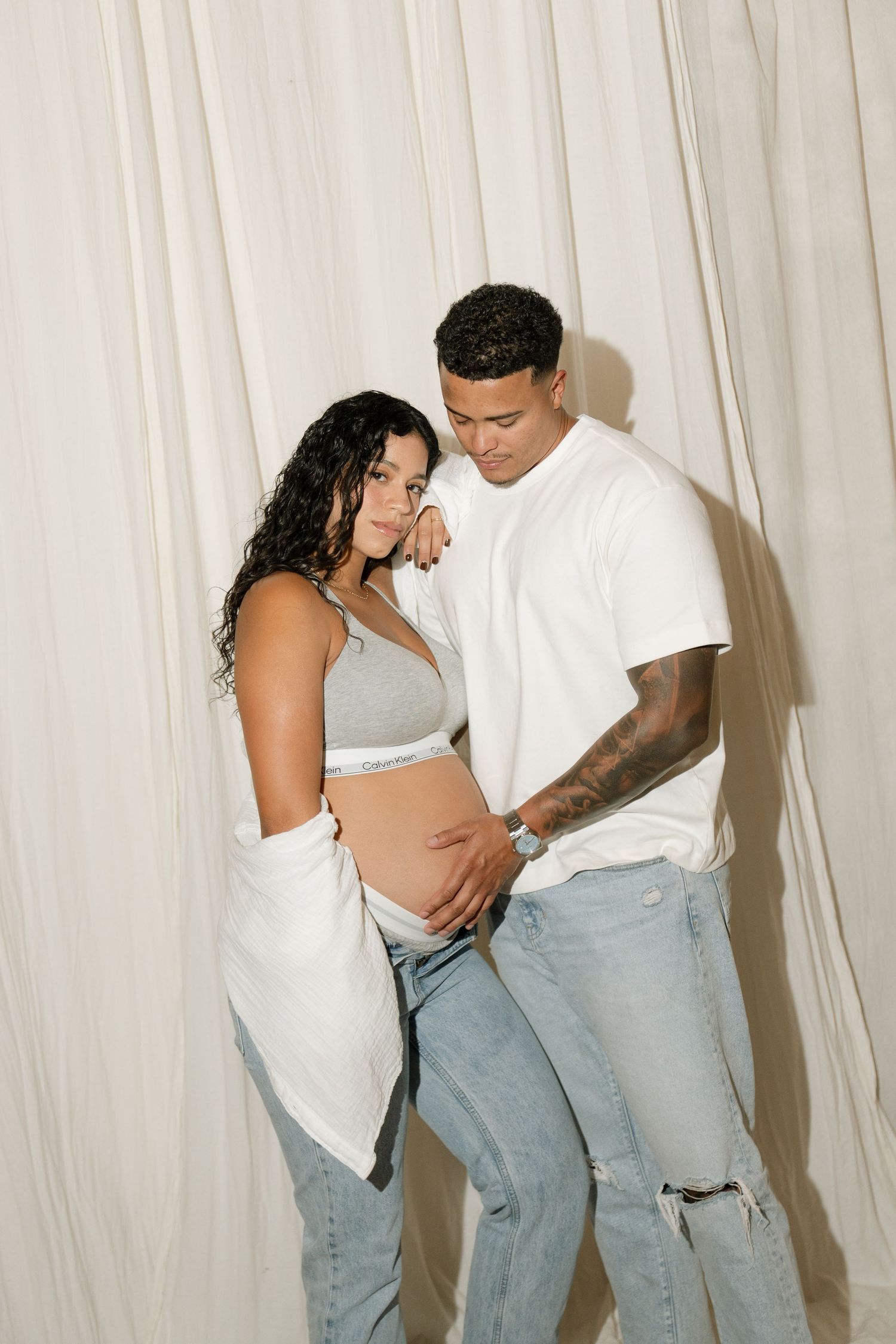 A couple poses against white drapes, woman in gray crop top showing baby bump, man in white tee tenderly touching her belly.