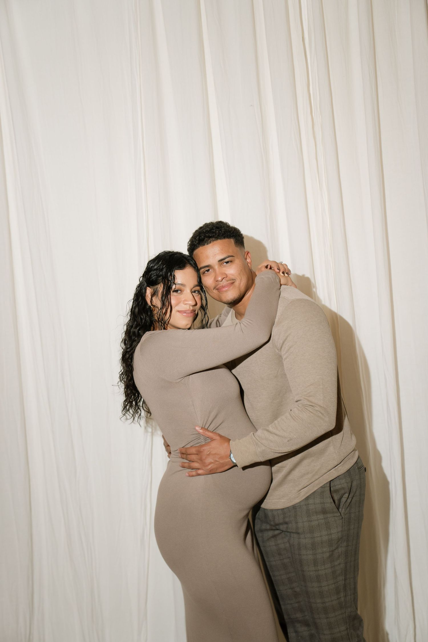 A couple embraces warmly against a white draped backdrop, both wearing coordinated beige outfits, smiling at the camera.
