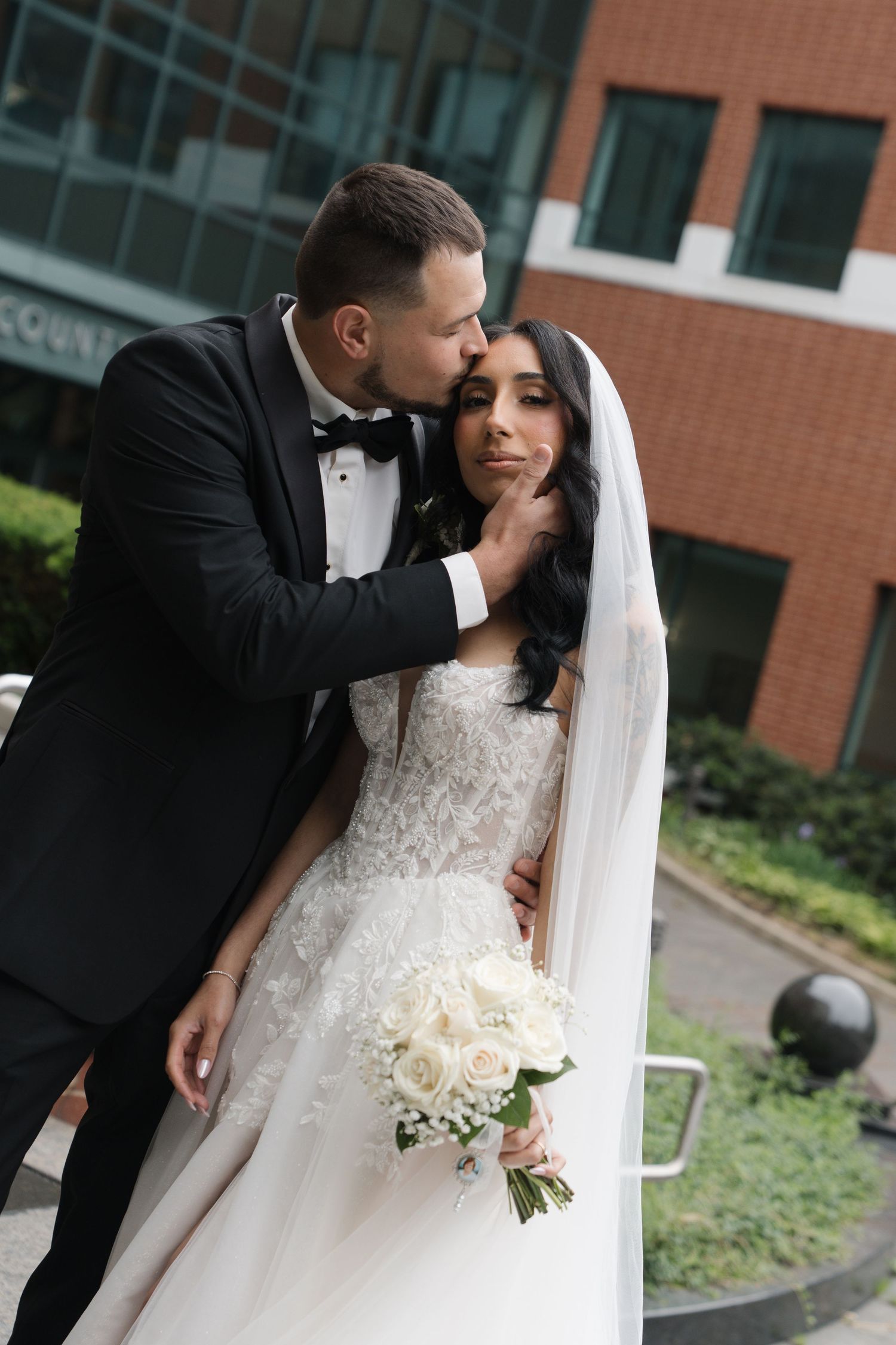 Groom in black tuxedo kissing bride in lace wedding dress holding white rose bouquet outdoors near brick building.
