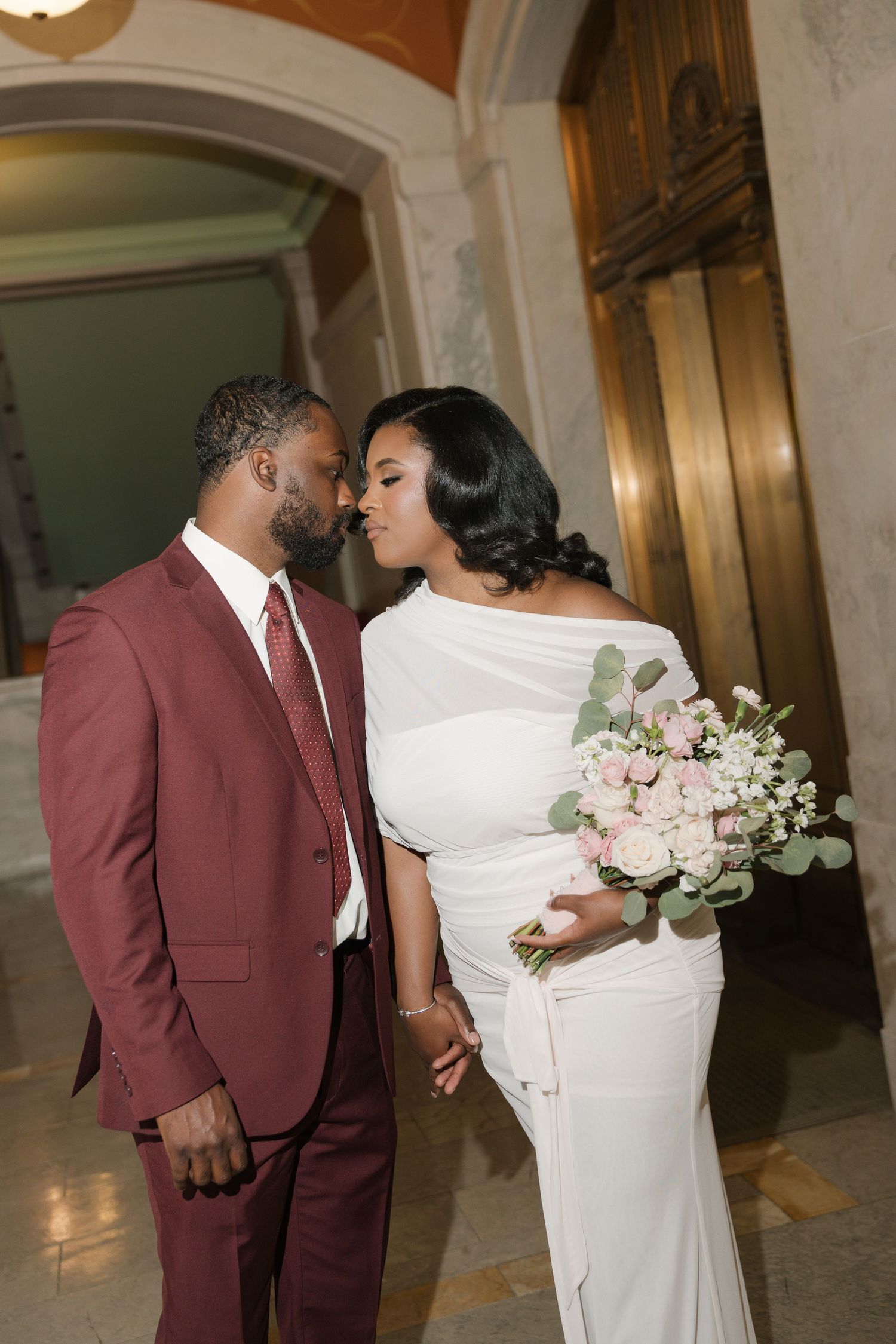 Couple in wedding attire share a romantic moment, groom in burgundy suit, bride in white gown holding pink roses bouquet.