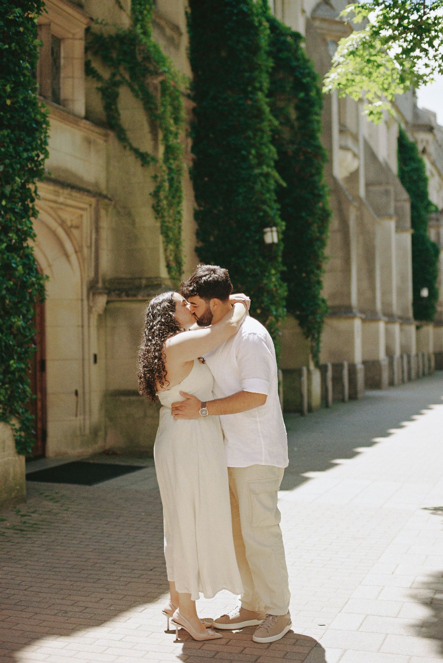 Couple sharing a romantic kiss outdoors beside an ivy-covered historic building on a sunny cobblestone street.