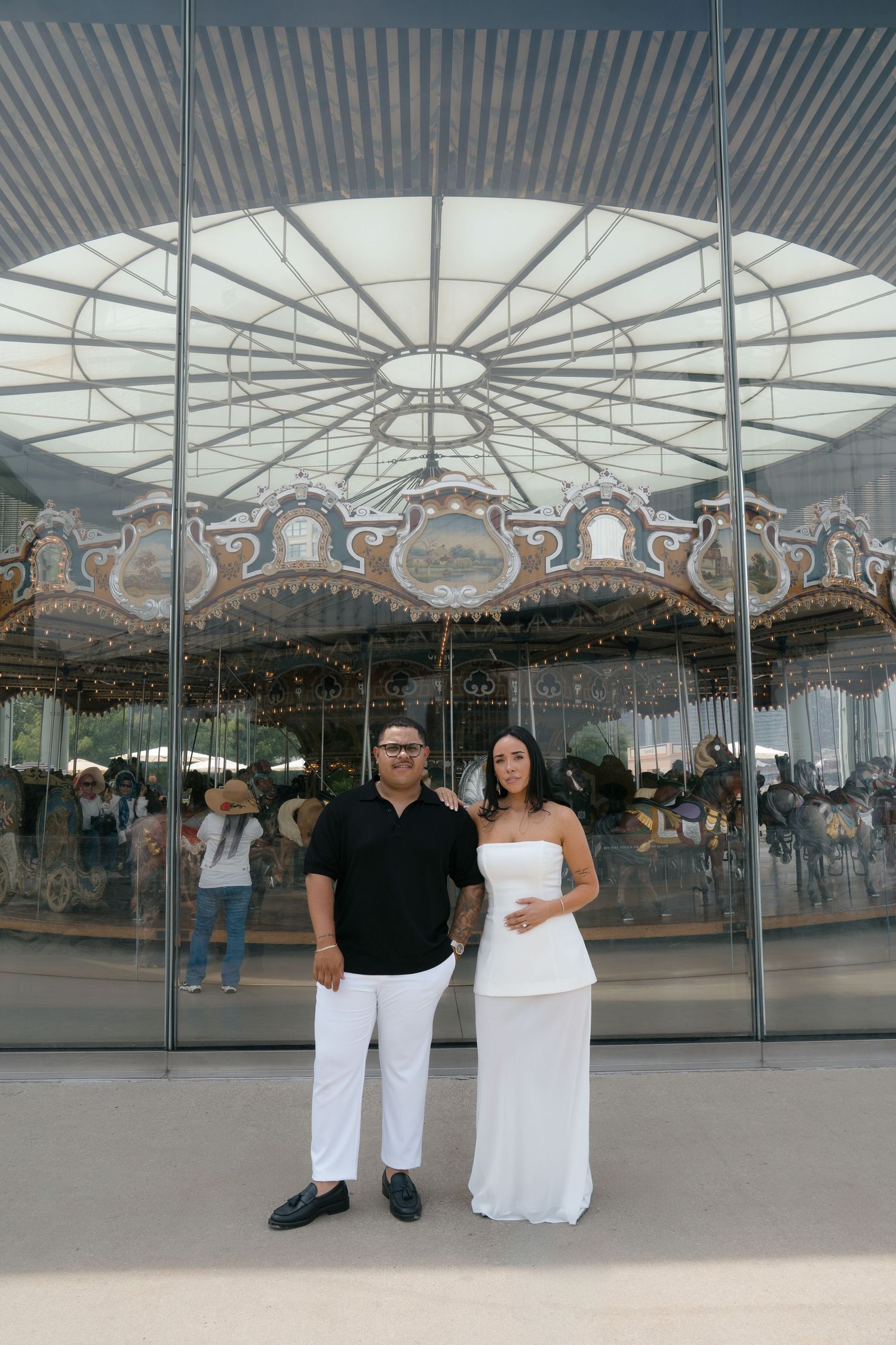 Couple in black and white outfits posing in front of an ornate carousel with decorative horses and lights.