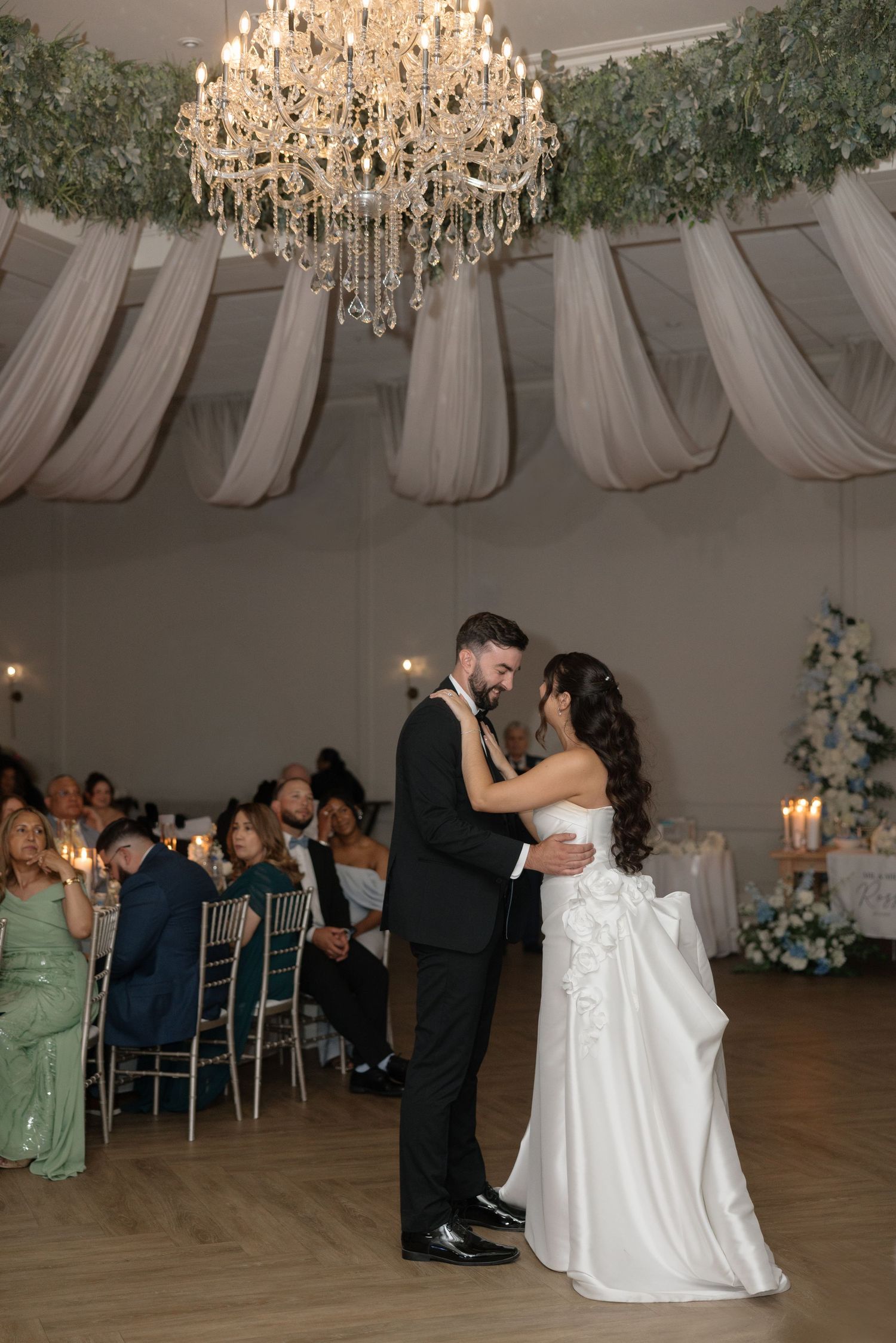 Bride and groom share first dance under crystal chandelier in elegant white draped reception hall.