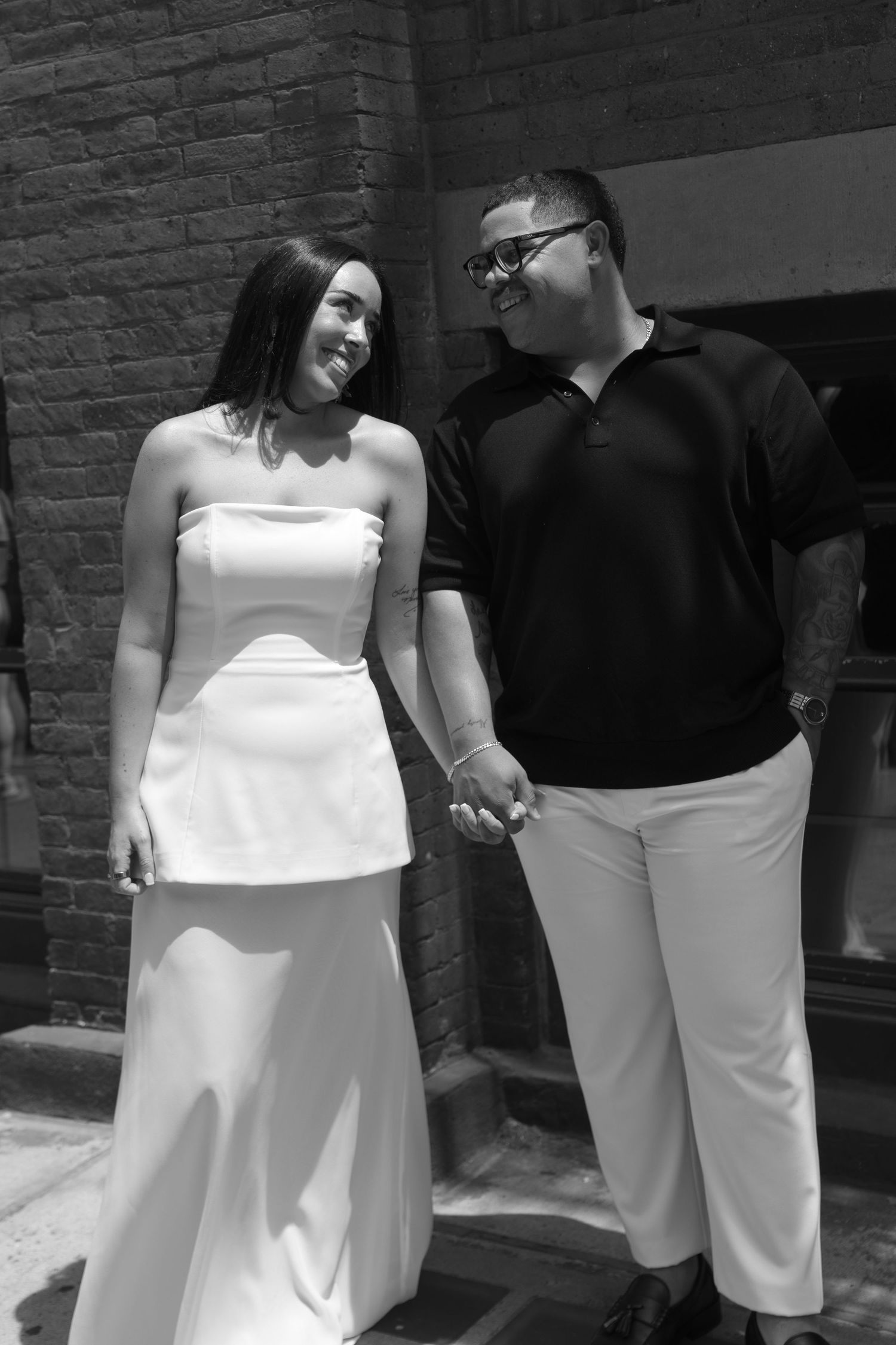 Black and white photo of a smiling couple holding hands, woman in white strapless gown, man in black polo and white pants.