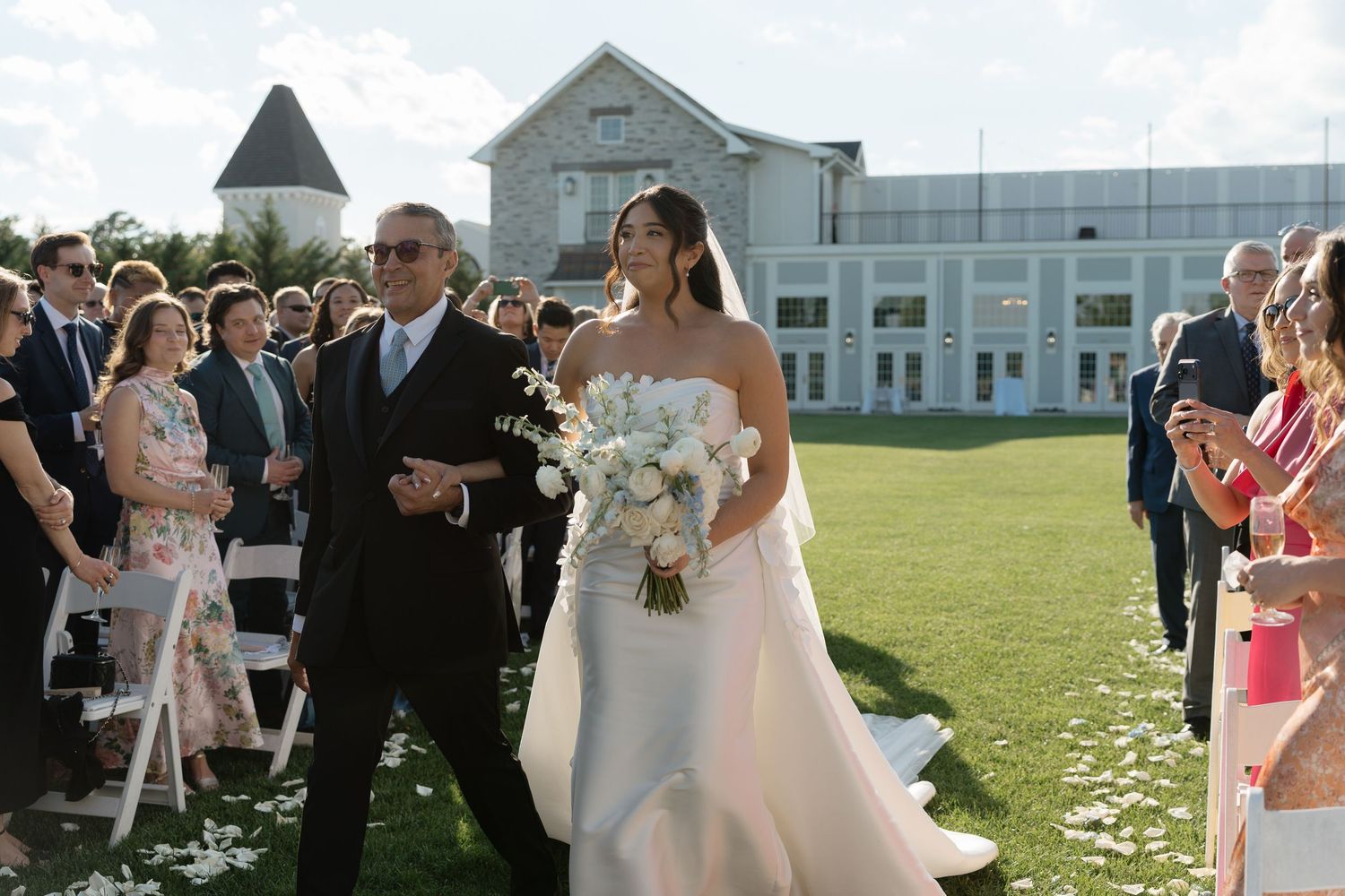 Bride in white gown walking down outdoor aisle with father on sunny wedding day.