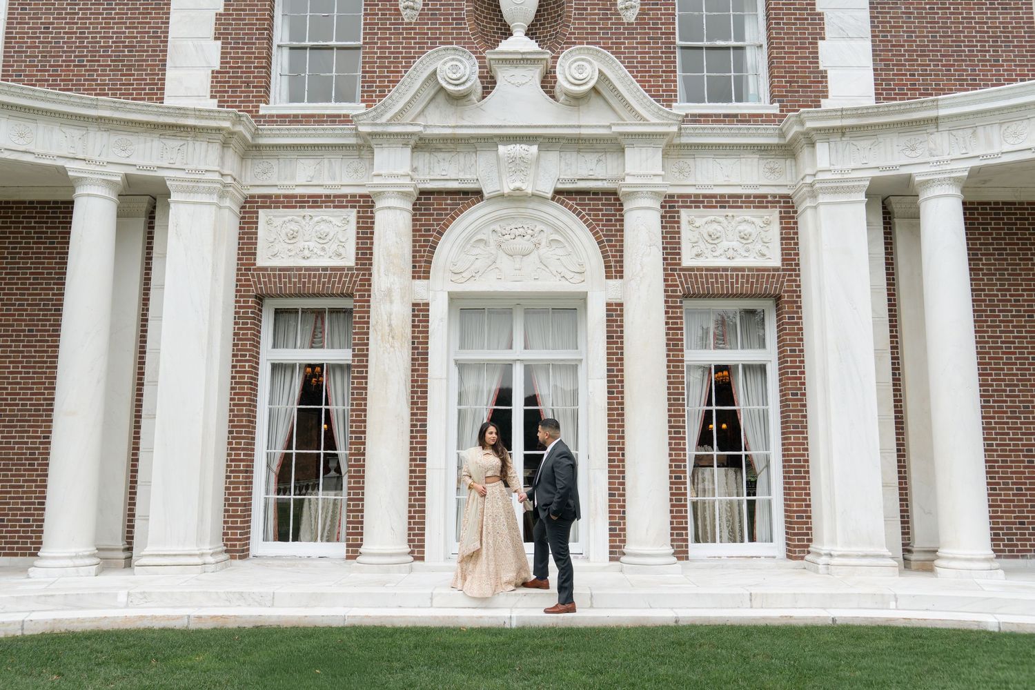 Elegant couple in formal attire stands in front of a grand colonial-style building with white columns and brick walls.
