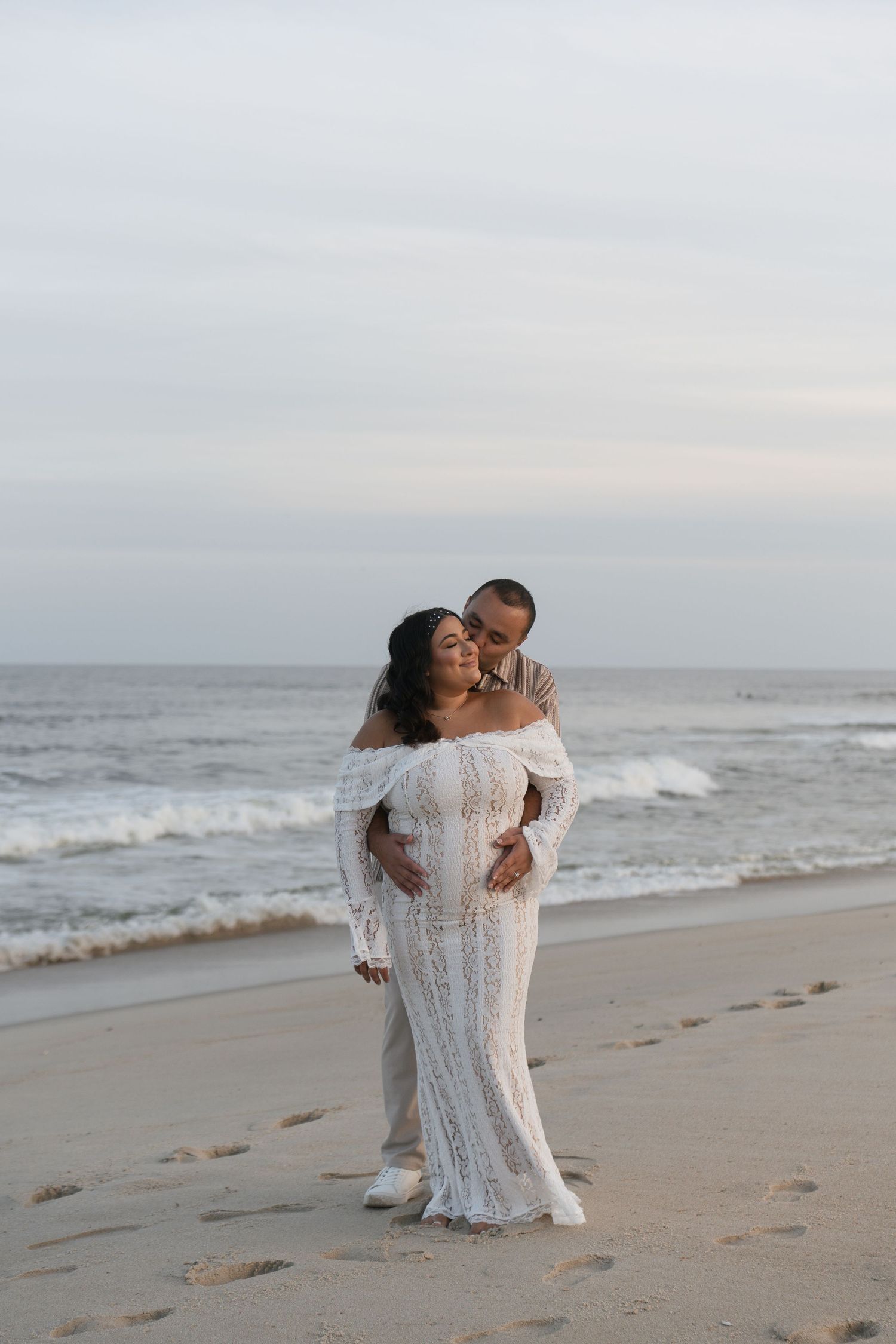 Couple sharing tender kiss on sandy beach, woman in white lace maternity gown at sunset.