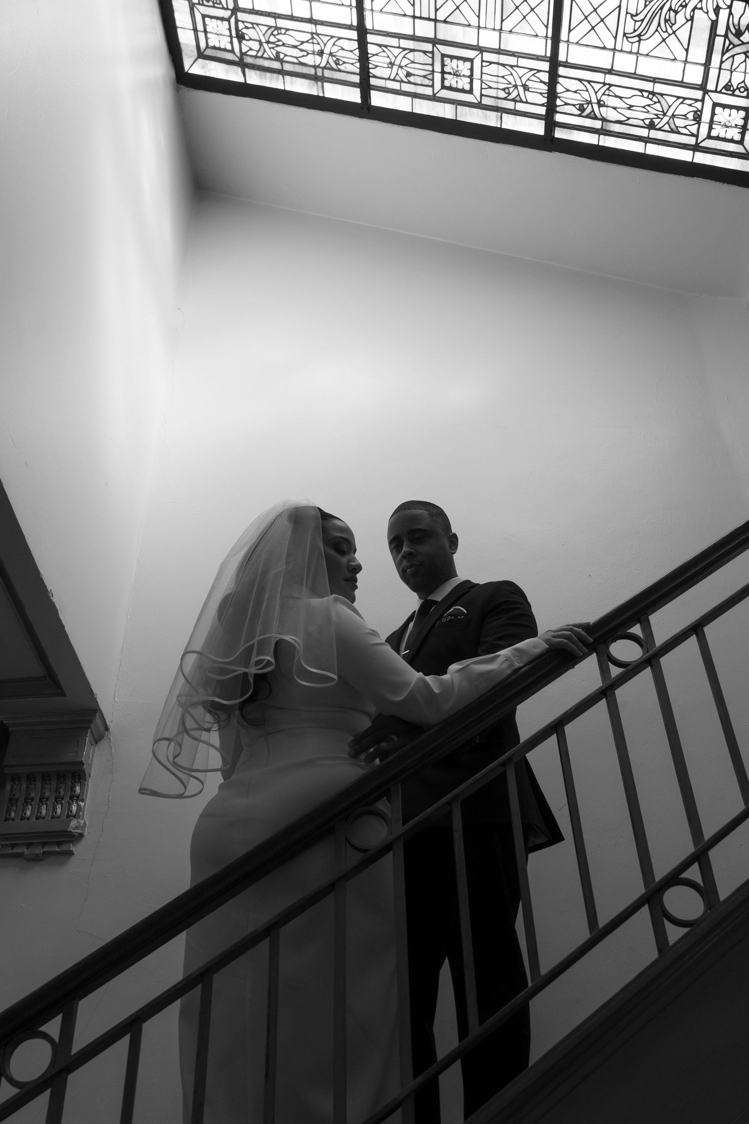 A black and white photo captures a couple on a staircase with decorative railings and window.