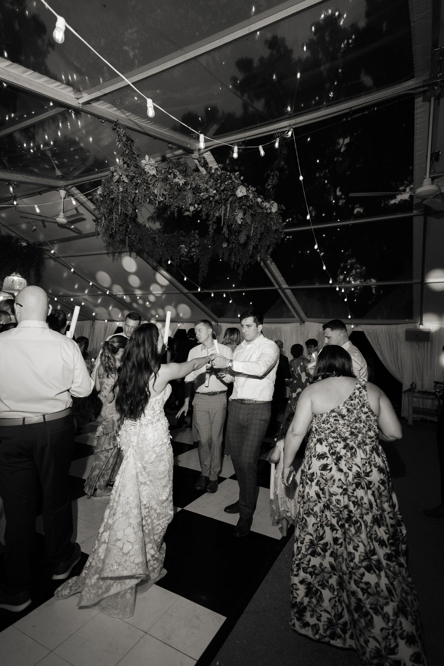 Wedding guests dance and celebrate under twinkling lights in a tented reception venue at night.