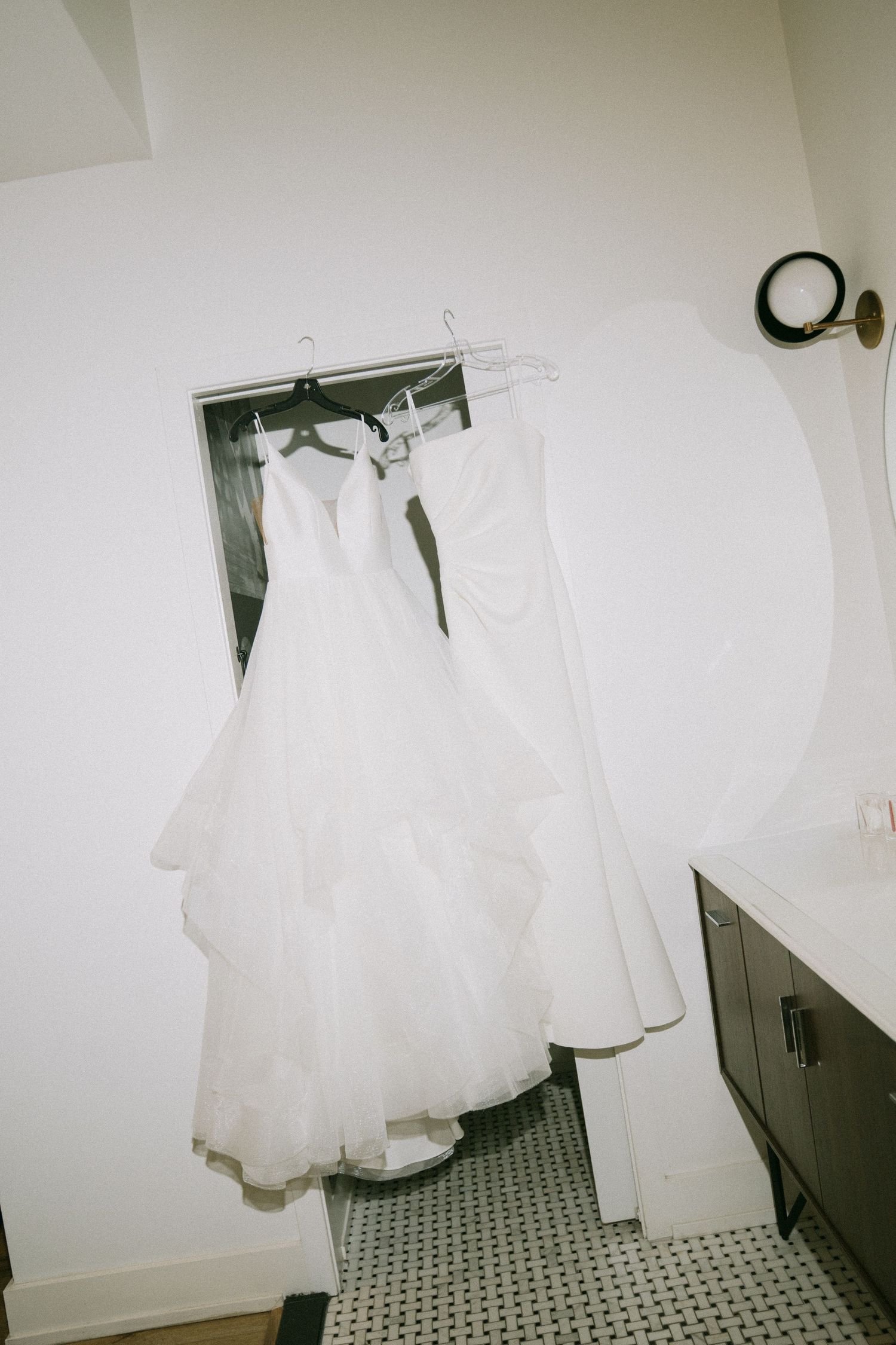A white wedding dress hangs in a bathroom doorway above black and white tile flooring.