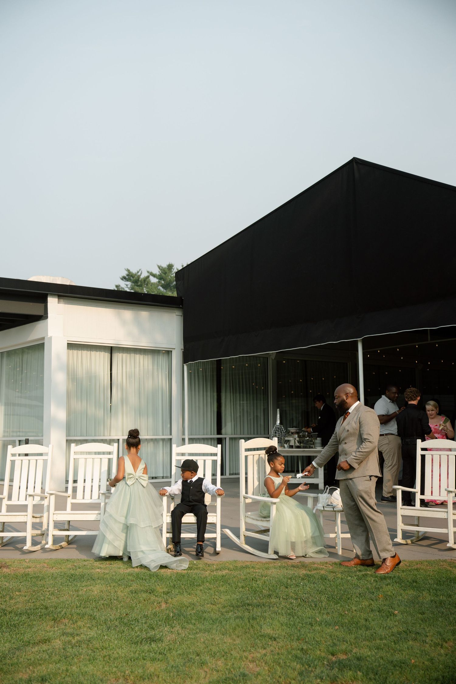 Wedding party with bride, groom, flower girls, and ring bearer gathered outside an elegant venue on a green lawn.