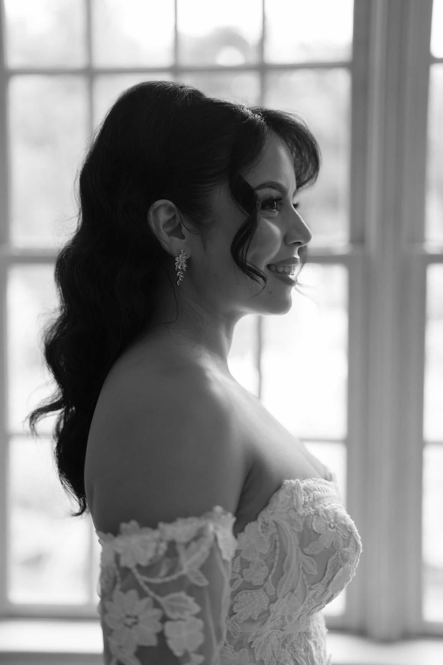 Black and white portrait of a smiling bride in an off-shoulder lace gown with long wavy hair by a window.